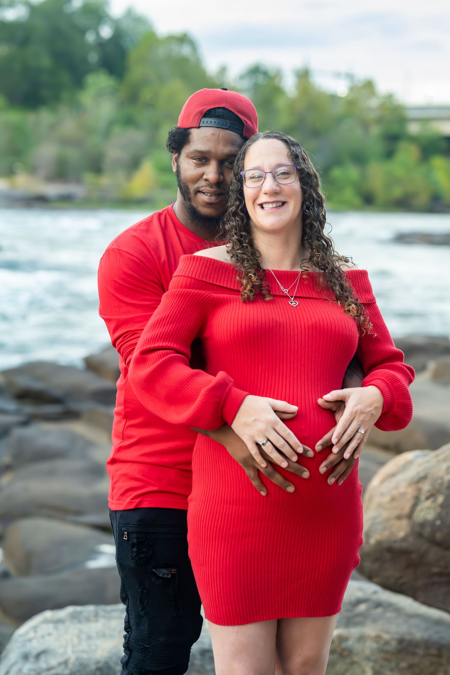 Couple in matching red outfits embrace on rocky shoreline during maternity photoshoot.