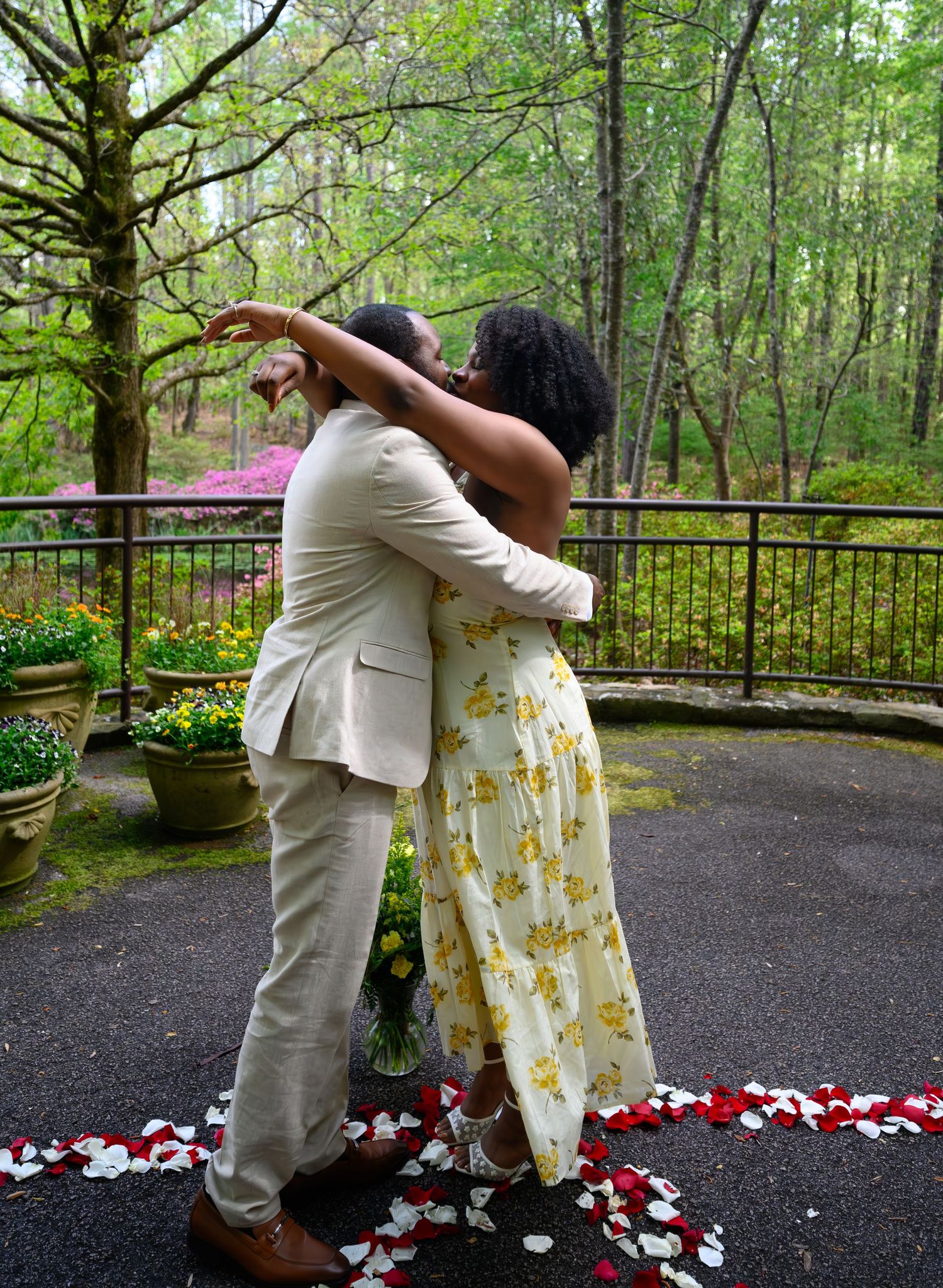 Couple embracing and dancing outdoors surrounded by rose petals on a garden terrace with lush green trees.