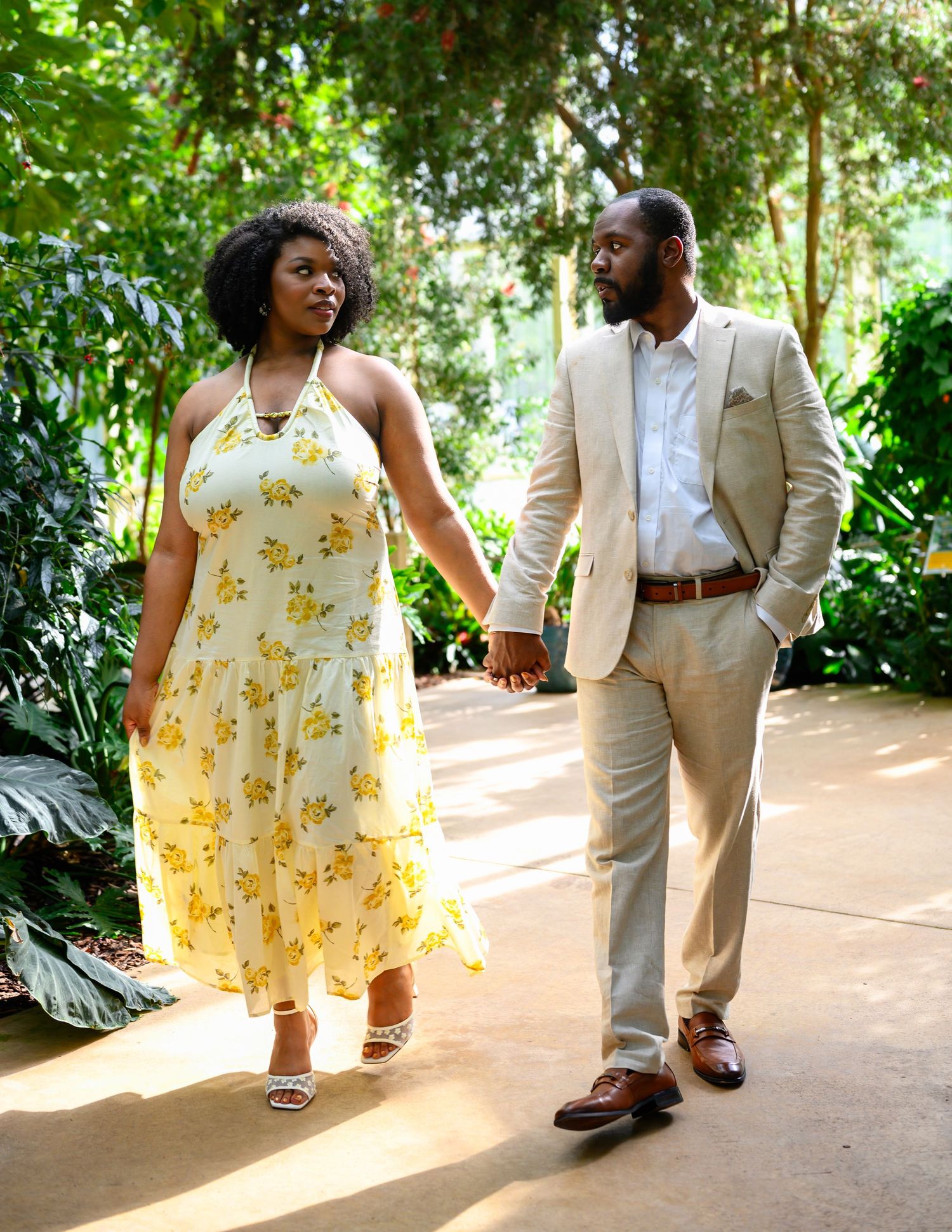 Engaged couple walking hand in hand through a lush tropical greenhouse, smiling at each other.