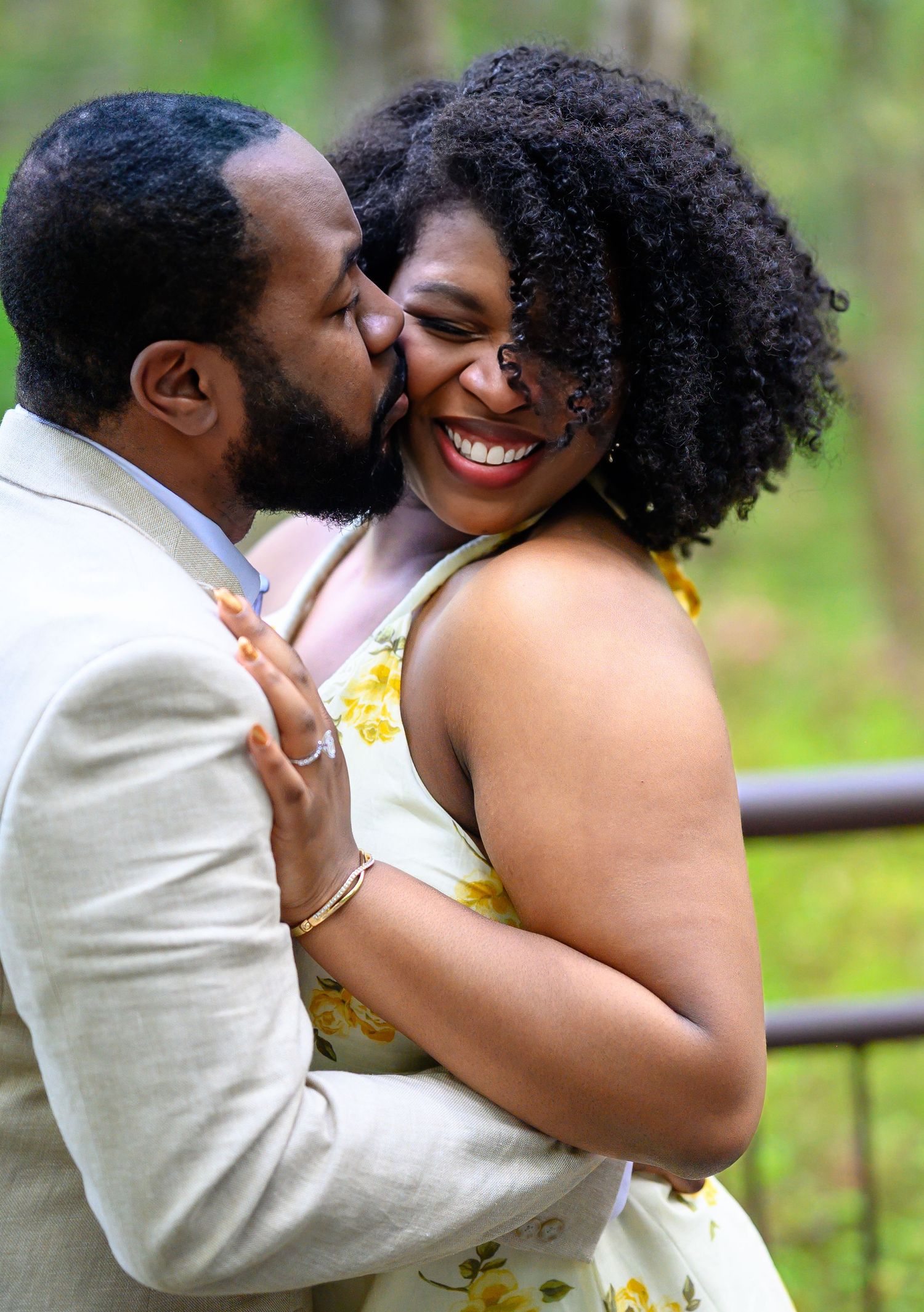 Man in beige suit kissing woman on cheek as she laughs joyfully outdoors in green wooded setting.