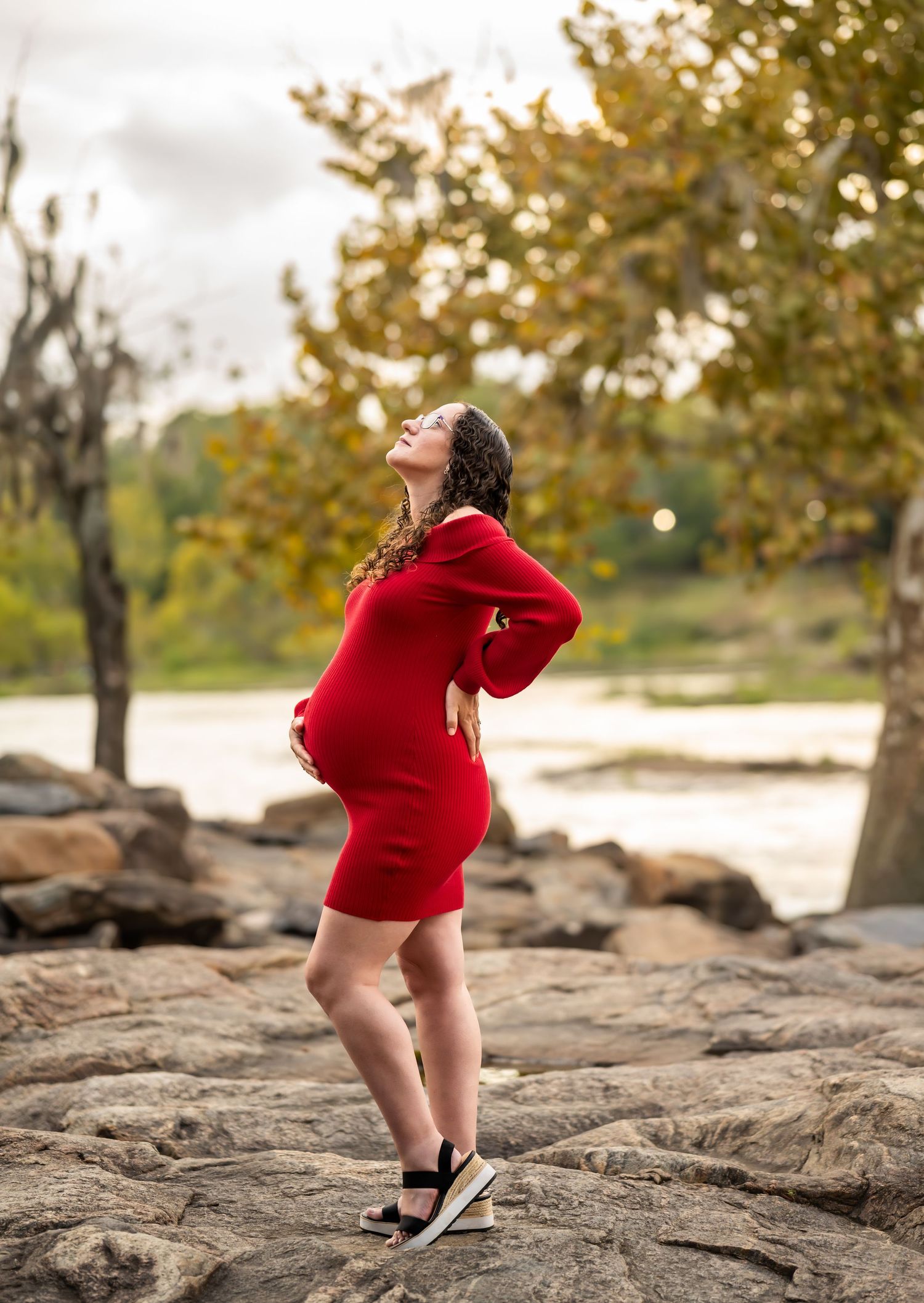 A person in a bright red sweater poses on rocky terrain near a waterfront.