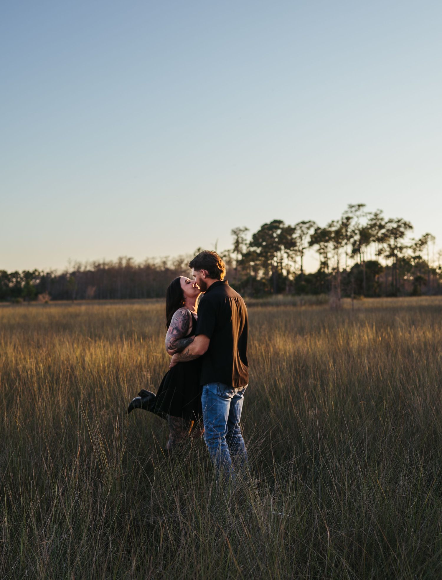 A couple embraces in a tall grass field during sunset with trees in the background.