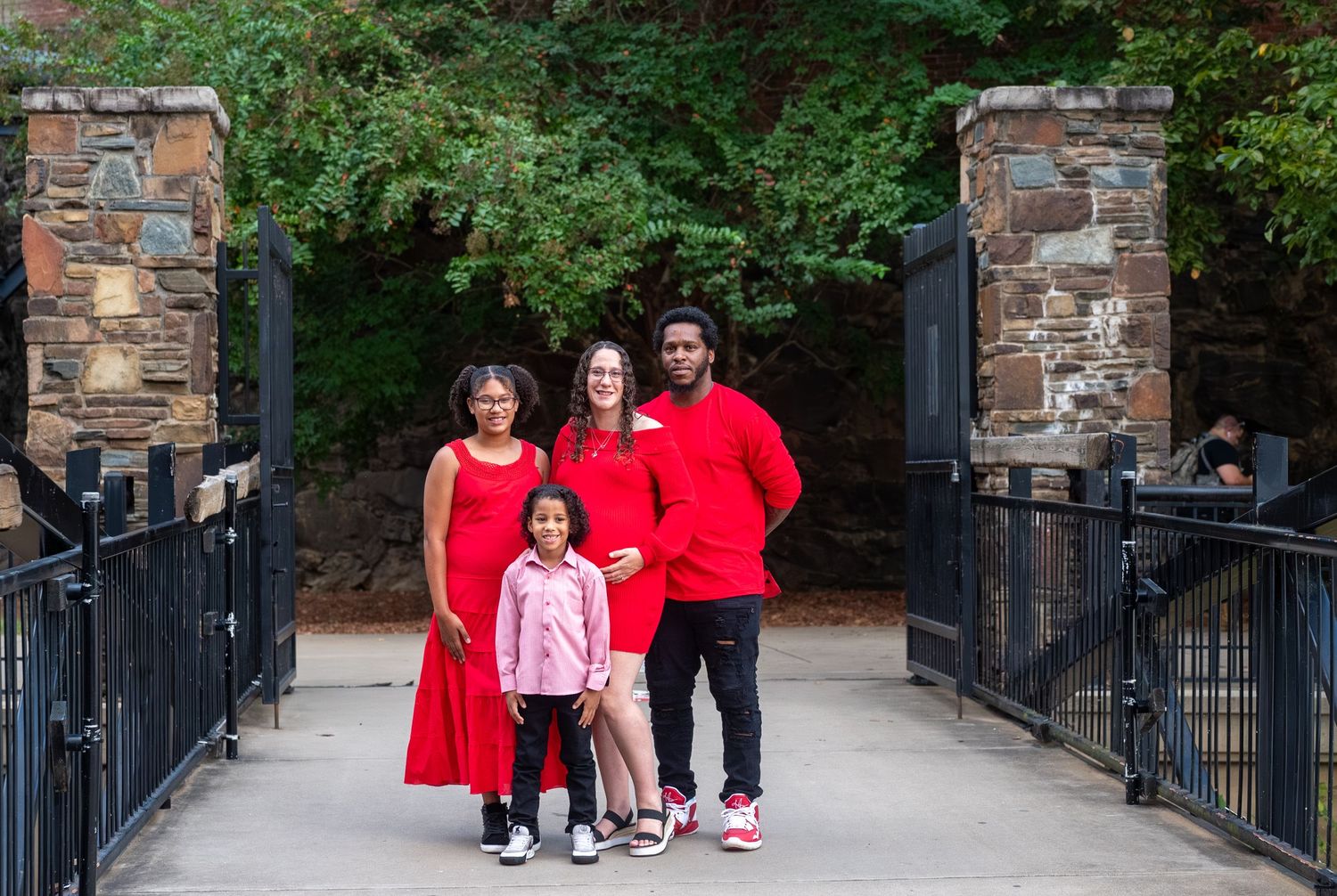 Family wearing coordinated red outfits stands together on stone bridge with black iron railings in park setting.