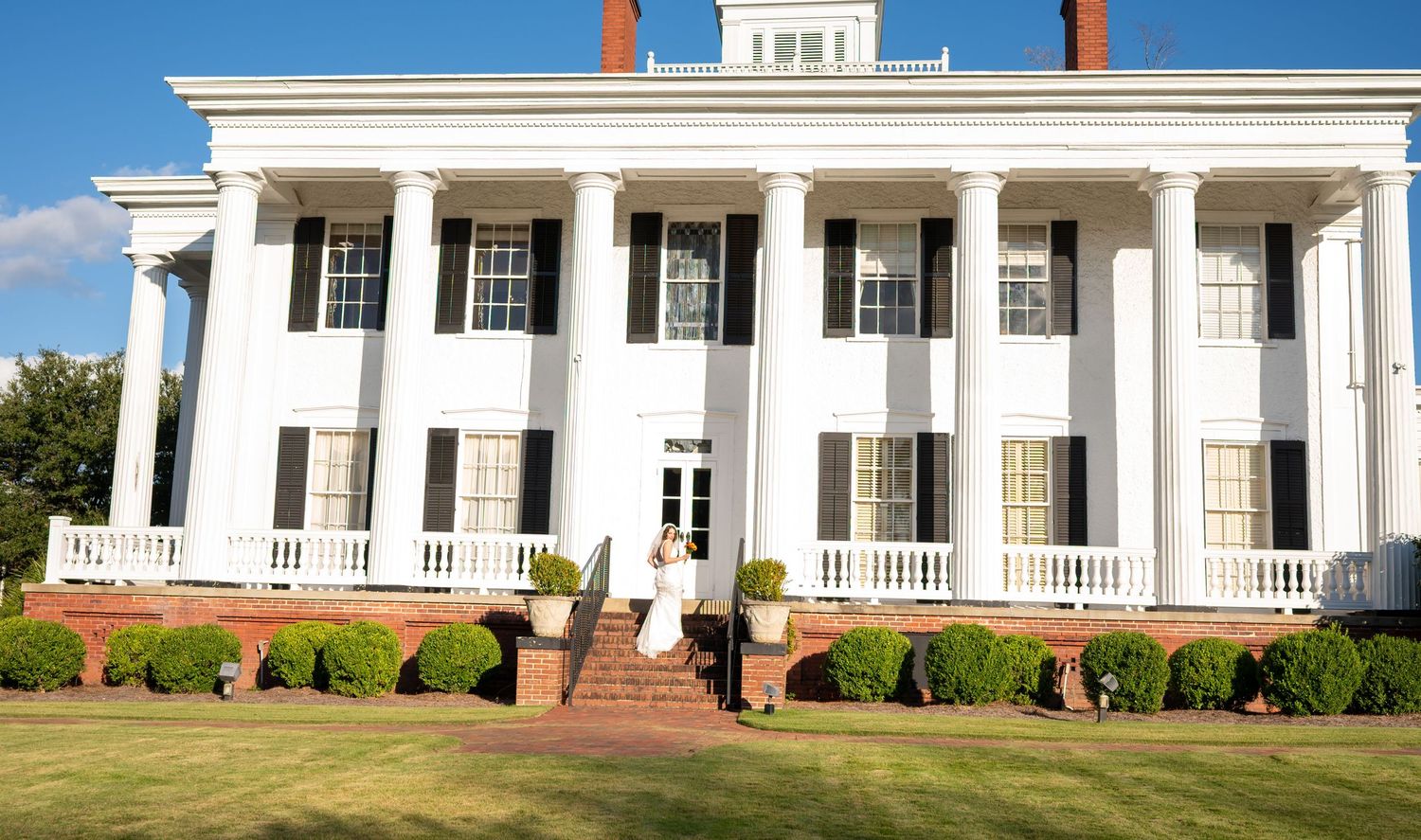 Historic white colonial mansion with tall pillars and wraparound porch features classic Southern antebellum architecture.