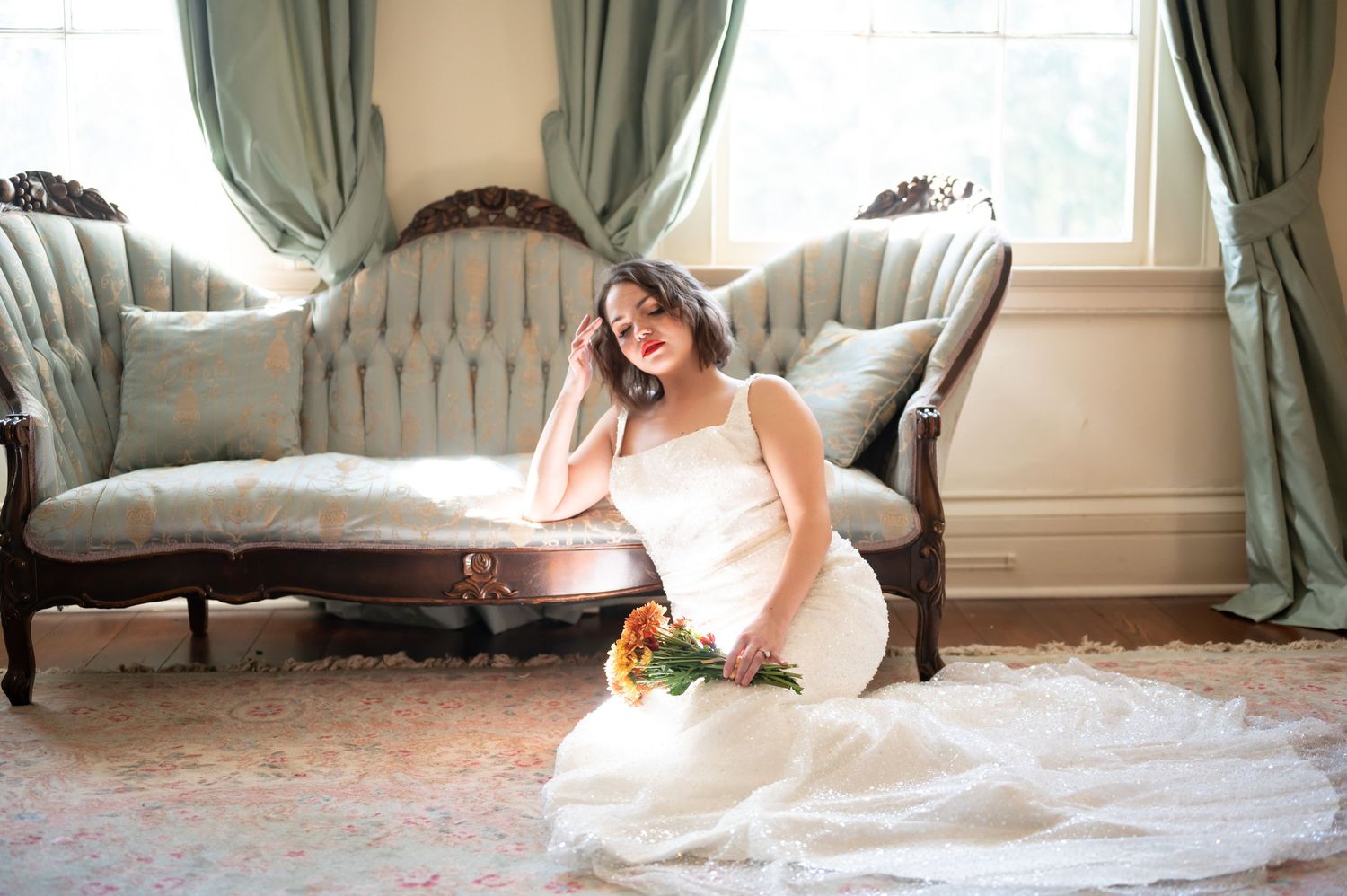 A bride in a flowing white wedding dress relaxes on a plush chaise lounge by a bright window.