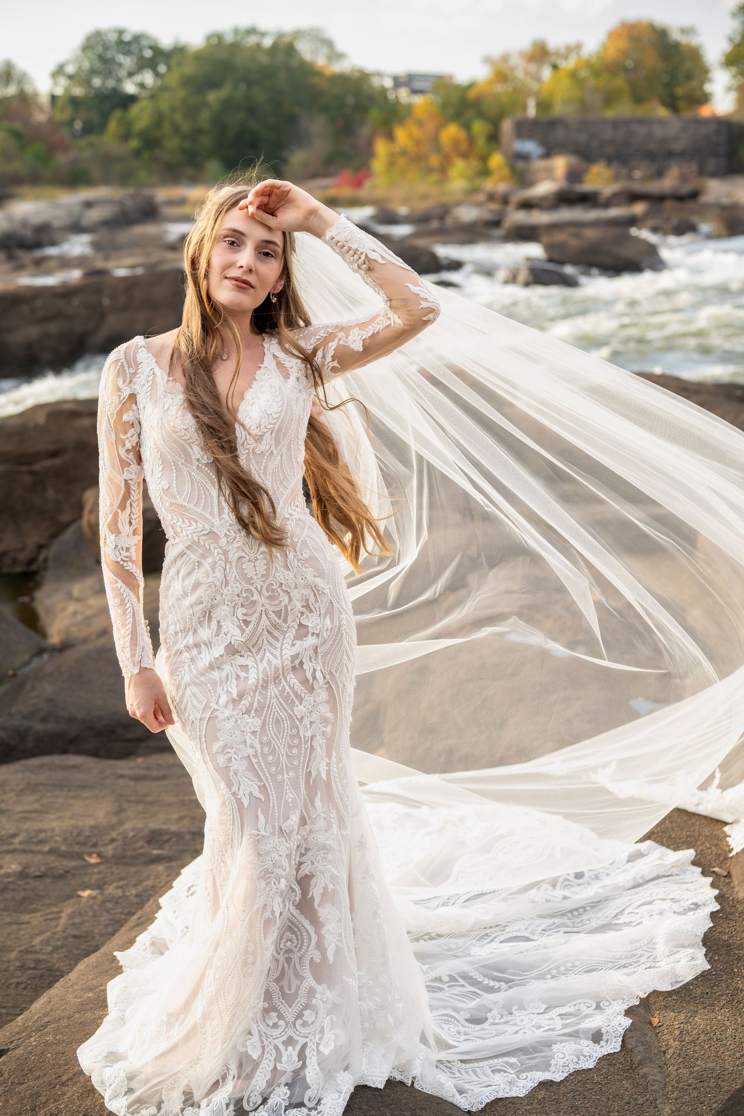 A flowing white lace wedding dress with long sleeves billows dramatically against a rocky riverbank in autumn.