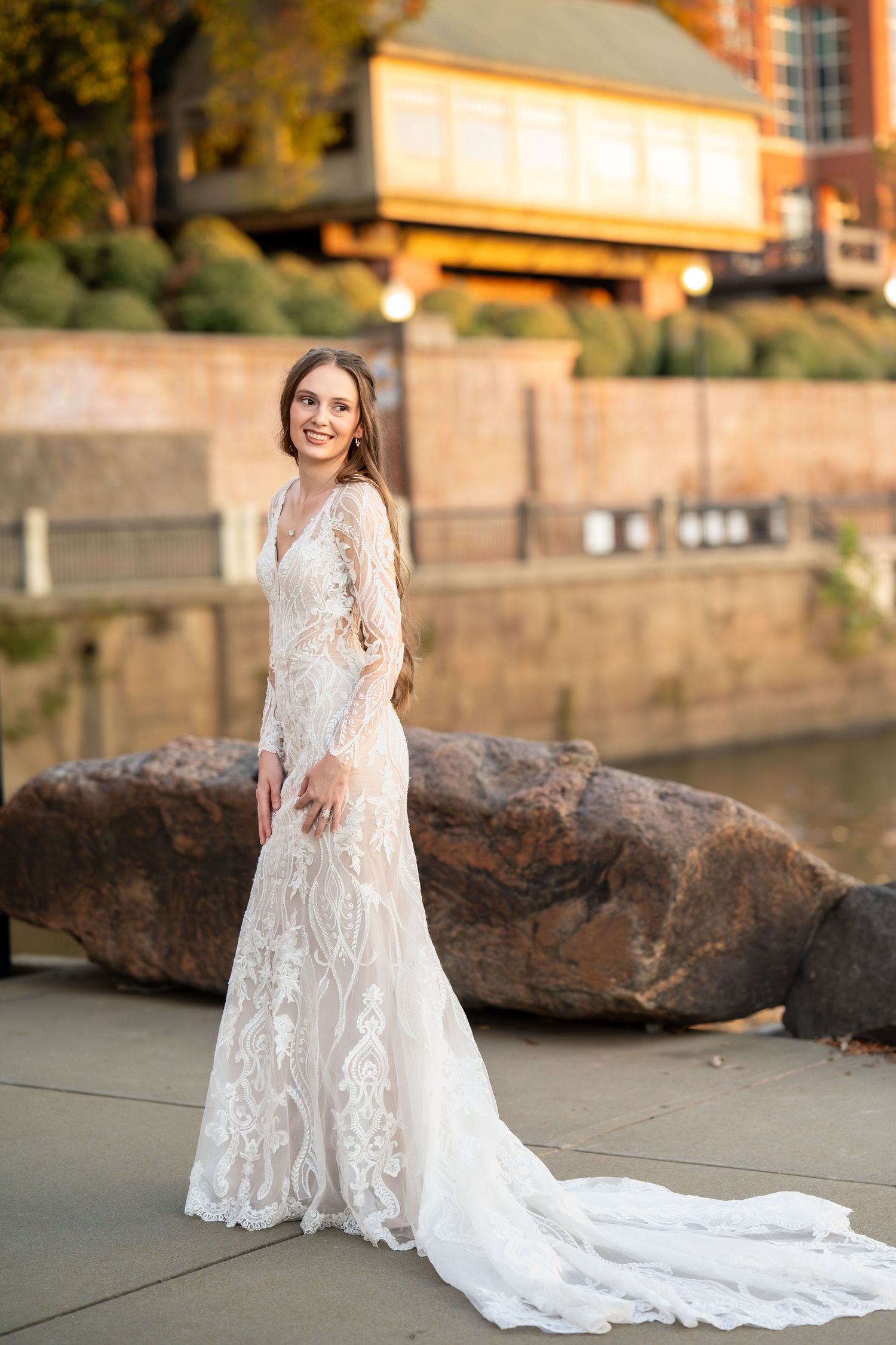 Bride in elegant lace gown with cathedral train poses by waterfront stone wall during autumn.