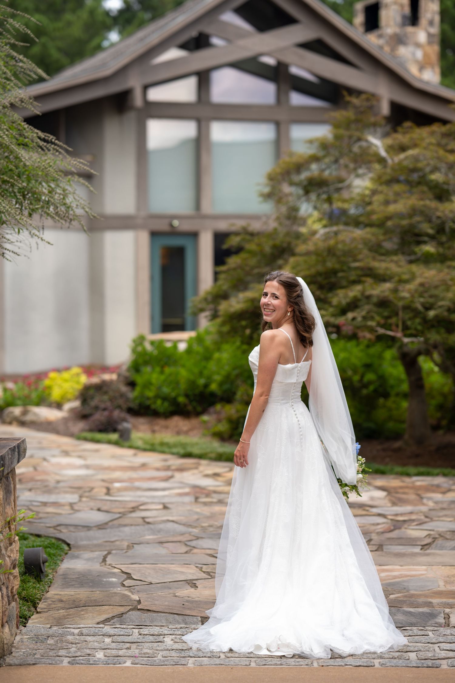 A bride in a flowing white wedding gown and veil poses on a stone patio outside a modern building.