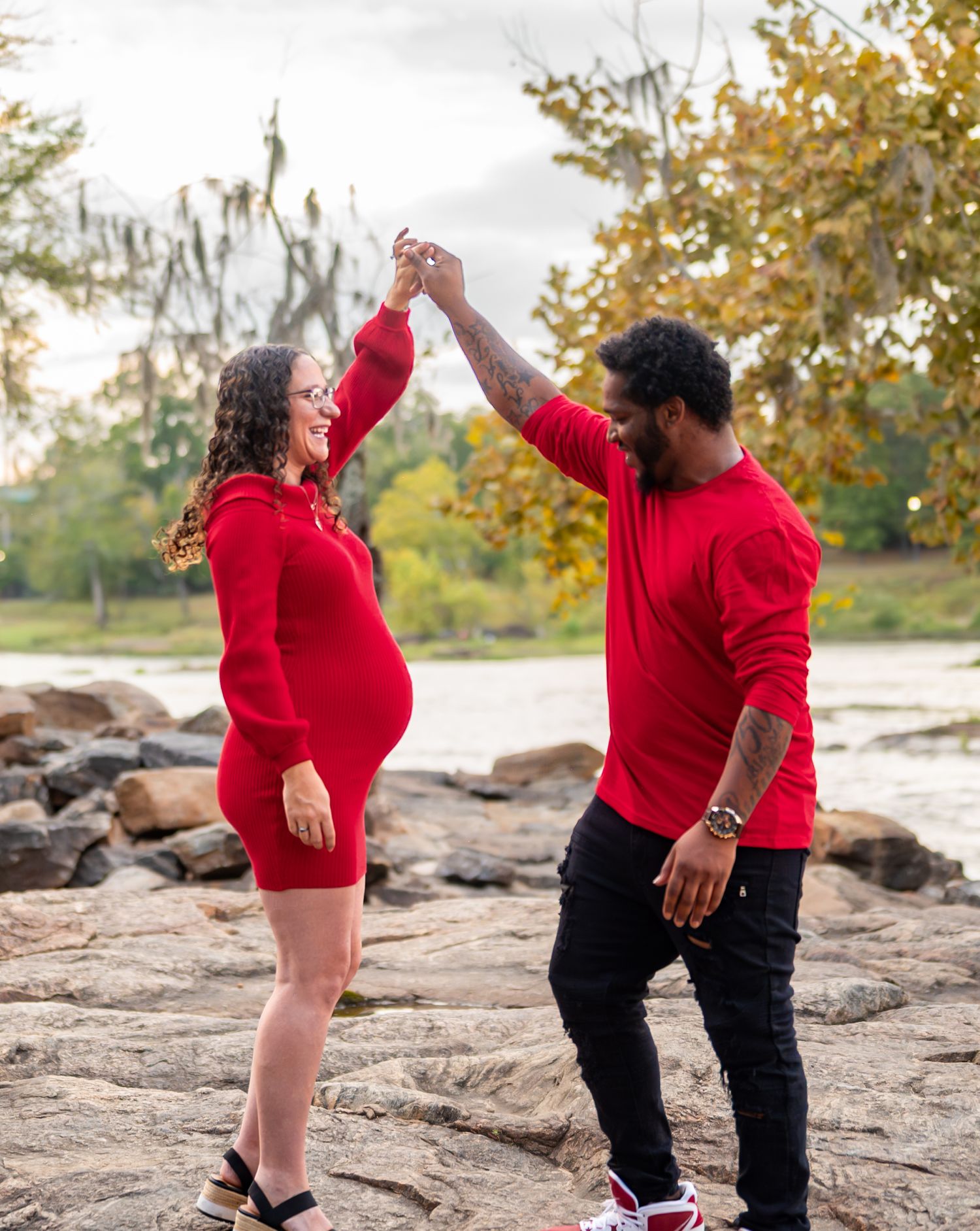 A couple in red attire strikes playful poses on rocky terrain by the water.