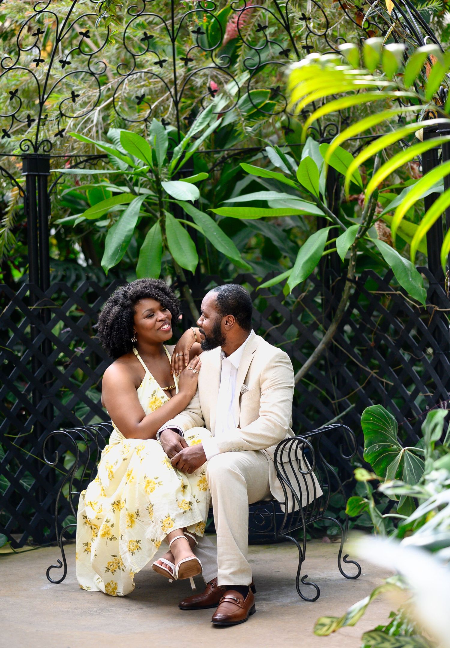 Newly engaged couple sitting on ornate bench surrounded by lush tropical garden greenery.