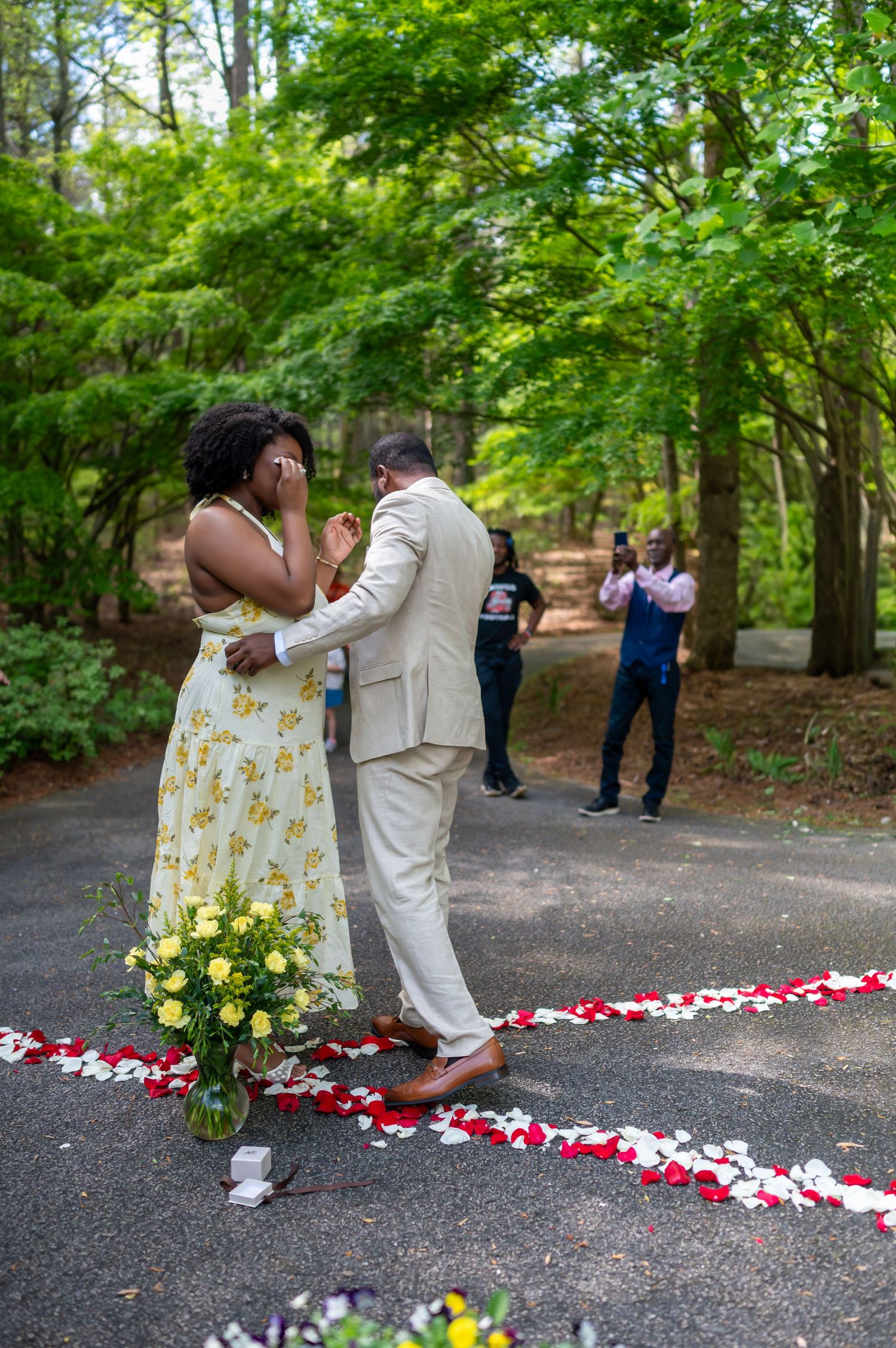 Emotional couple embraces during a romantic outdoor proposal surrounded by rose petals and yellow flowers in a forest setting.