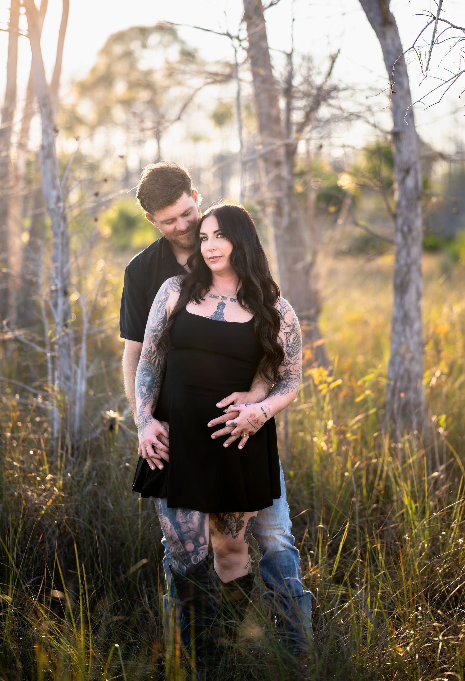 Romantic couple embraces in a sunlit forest with tall grass and birch trees during golden hour.
