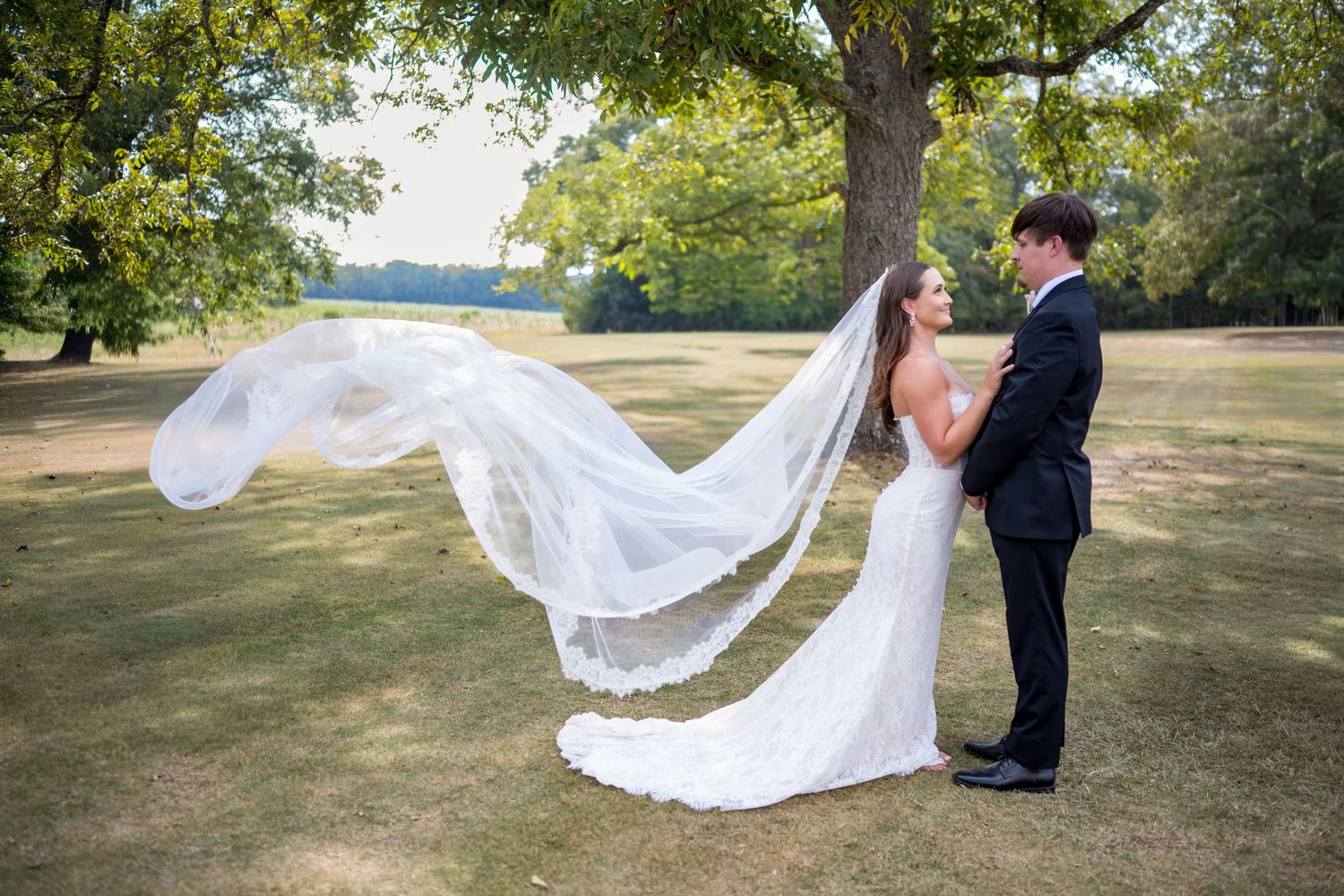 A couple in wedding attire stands together in a park as a flowing white veil catches the breeze behind them.