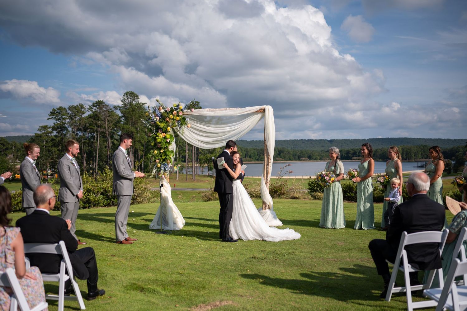 A scenic outdoor wedding ceremony takes place on a grassy hillside overlooking a lake under dramatic cloudy skies.