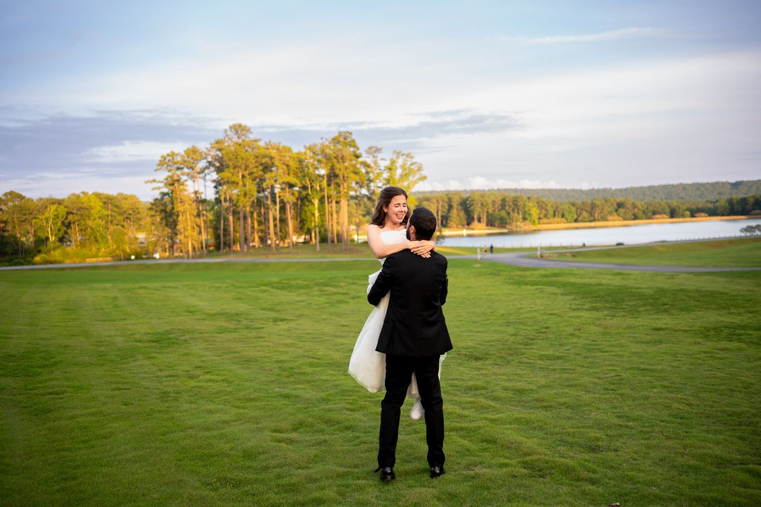 A couple walks hand in hand across a scenic grassy field with autumn trees in the background.