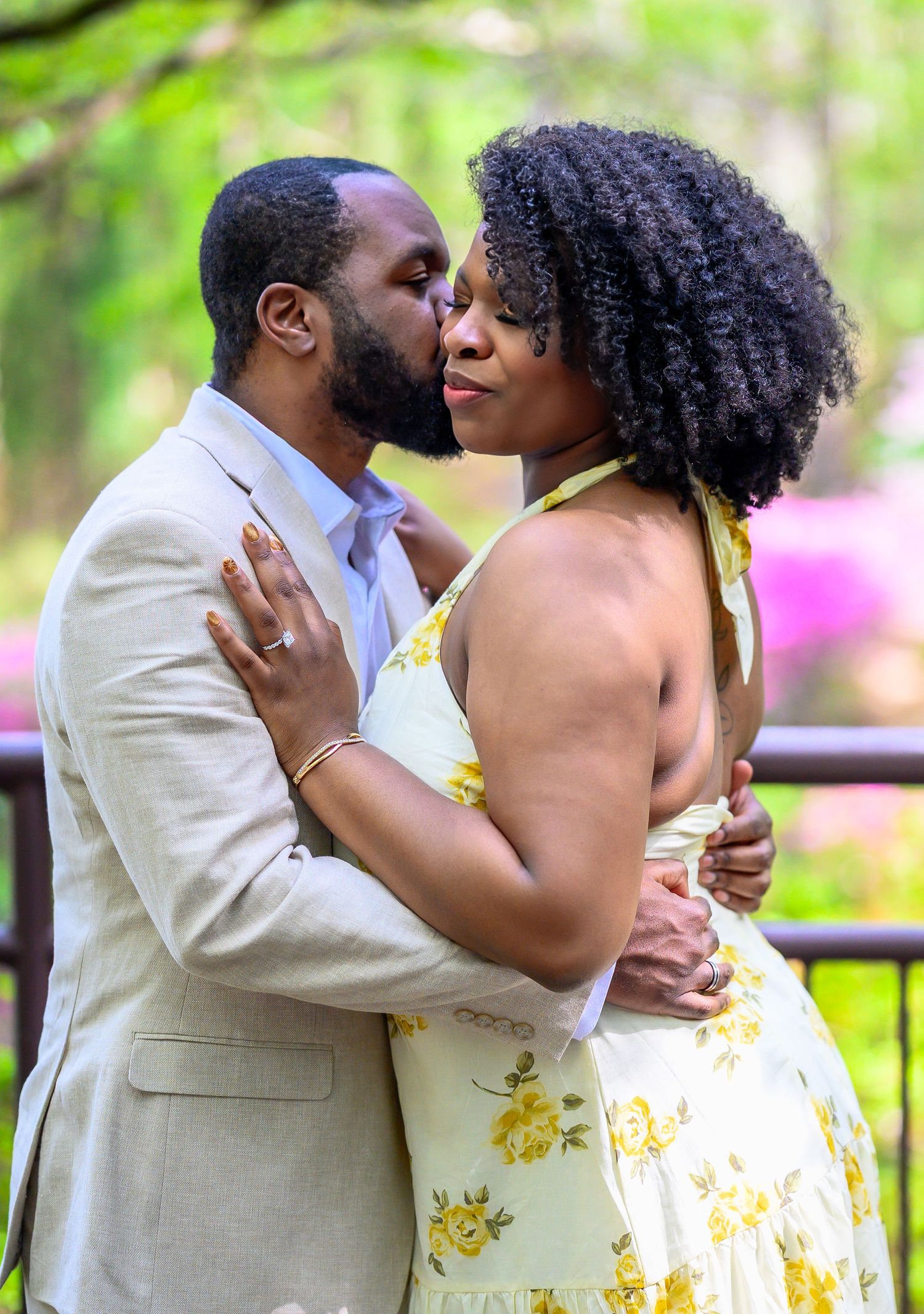Man tenderly kissing woman on the cheek outdoors with pink flowering bushes in the background.