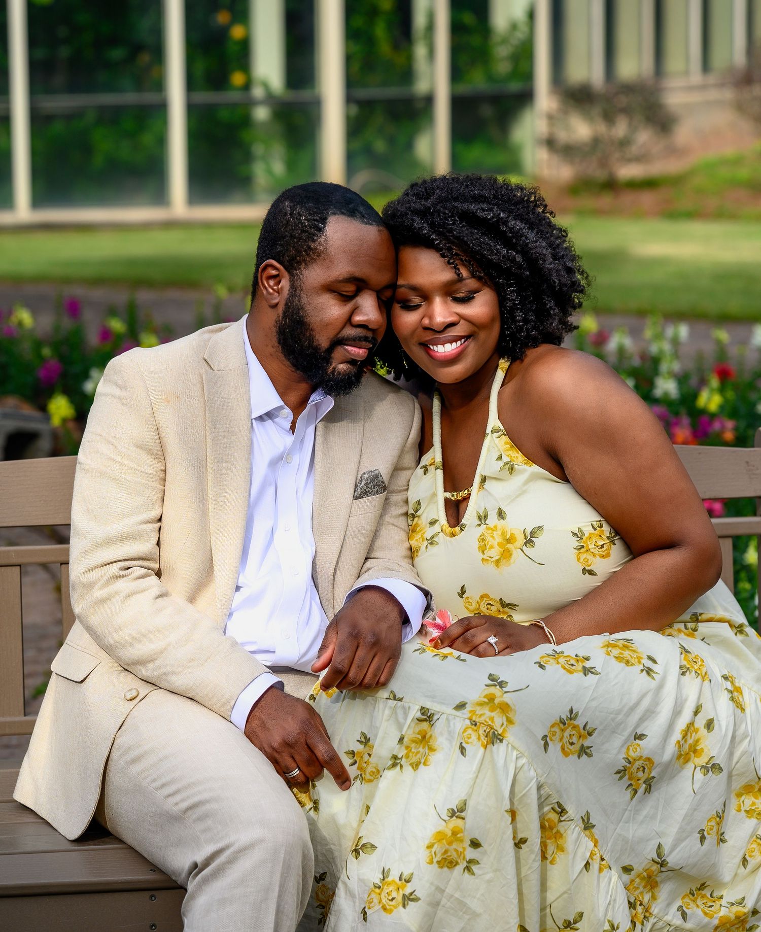 Couple sitting closely on a bench, smiling tenderly at each other in a vibrant flower garden setting.