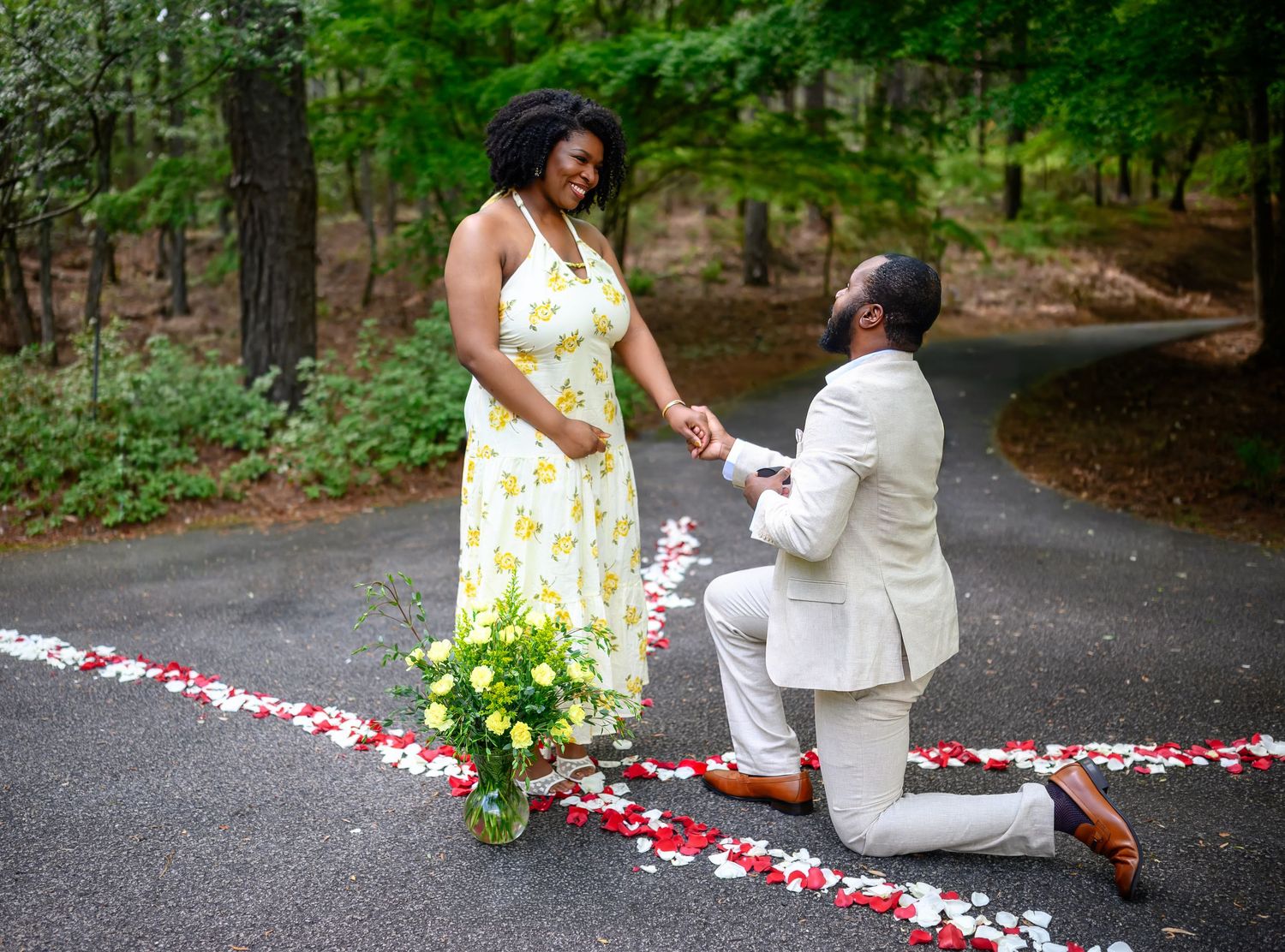 A man proposes on one knee to a woman on a petal-lined path with a yellow flower bouquet nearby.