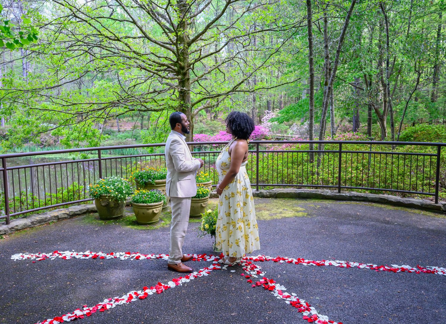 Couple stands facing each other on a patio surrounded by rose petals and lush greenery during a romantic proposal.