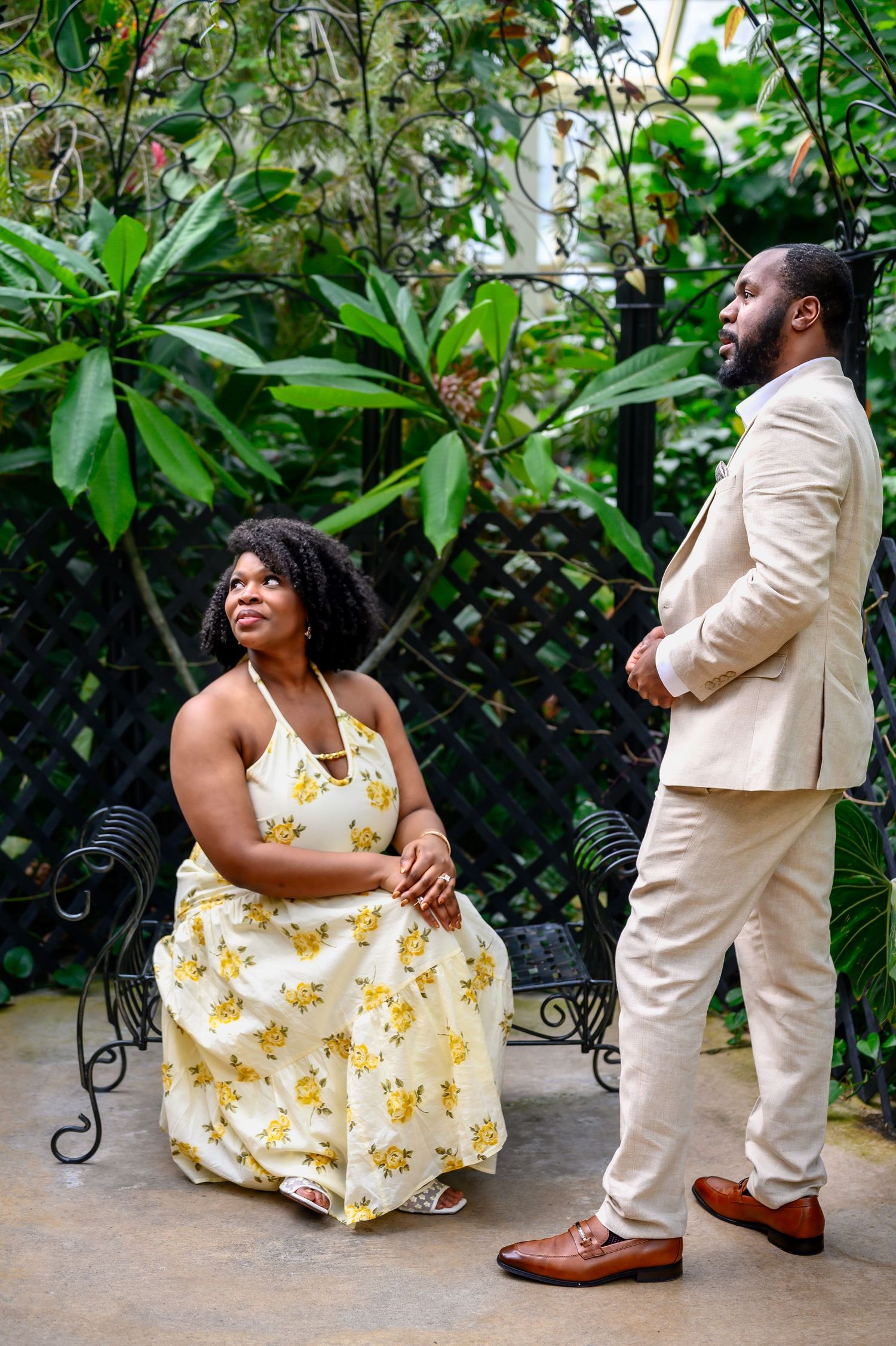 Couple sharing a tender moment in tropical garden, woman seated on bench in yellow floral dress looking up at man.