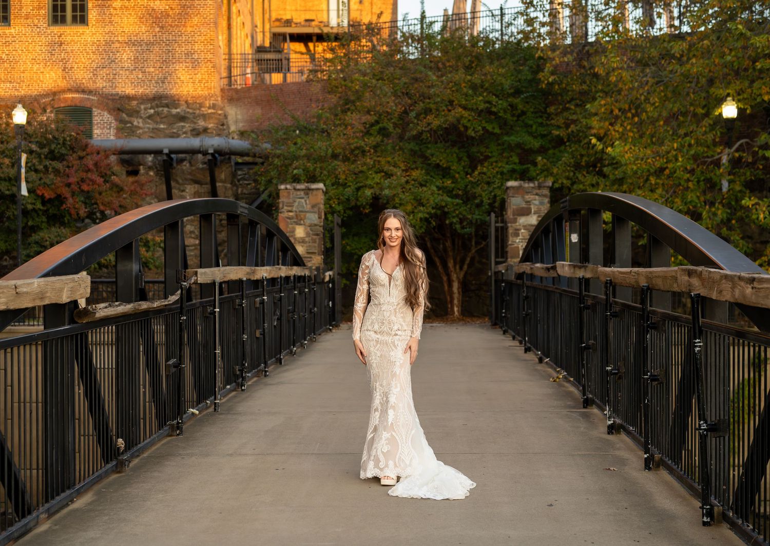 Bride in form-fitting lace gown stands centered on iron bridge with autumn foliage backdrop.