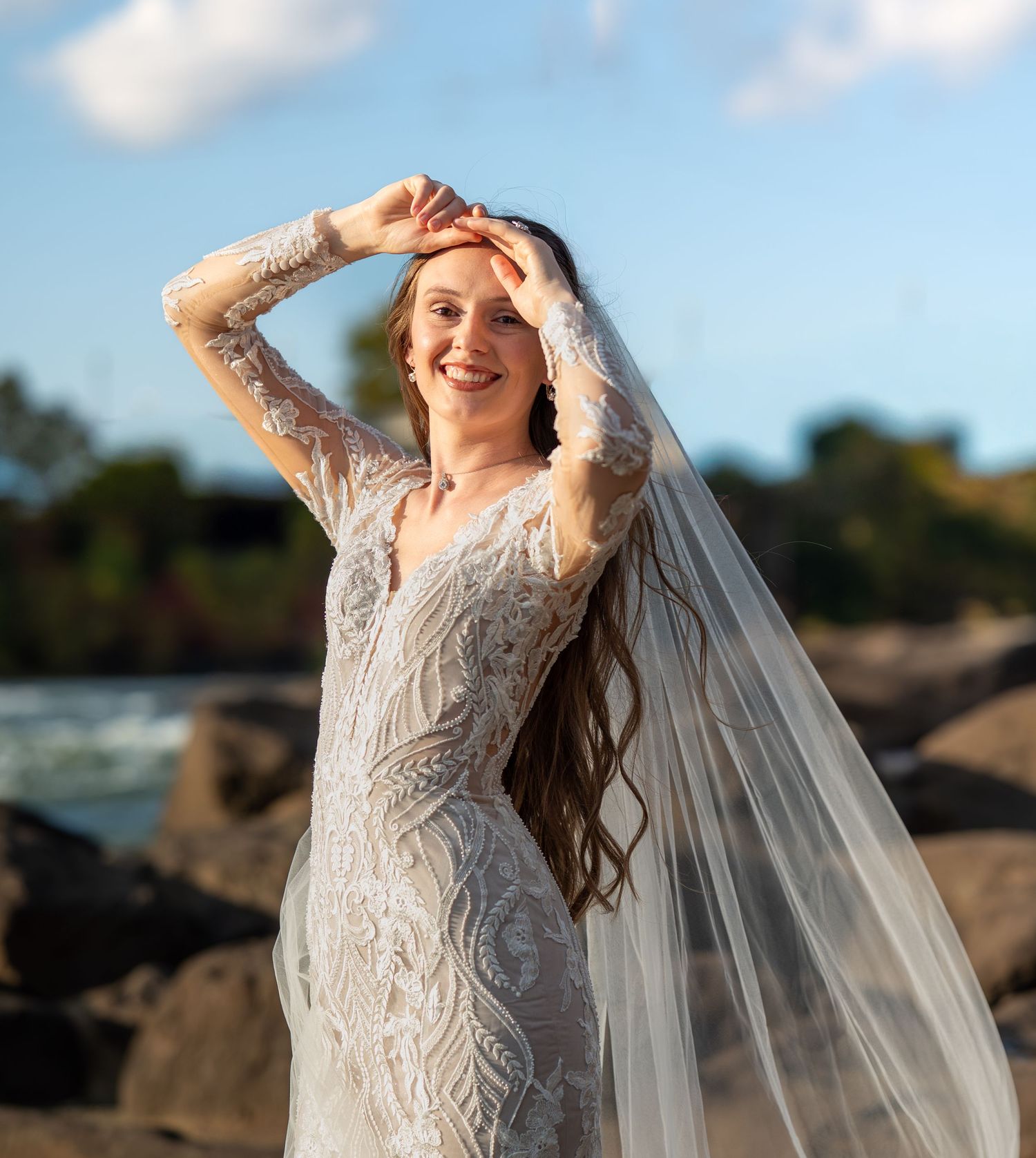 A bride in a detailed lace wedding dress poses dramatically against a rocky coastal backdrop at sunset.