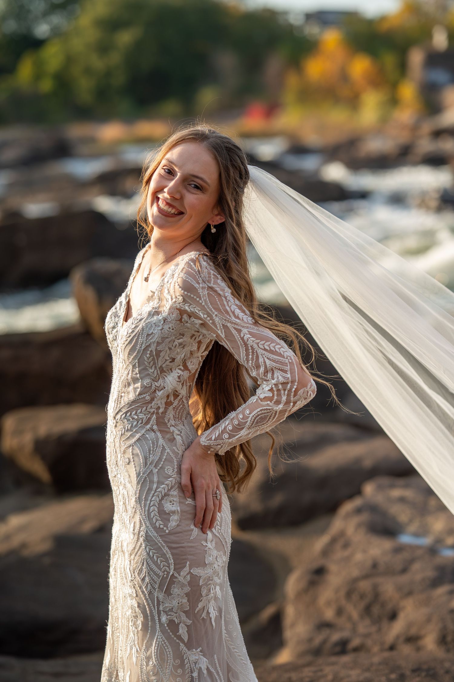 An elegant bridal portrait featuring a flowing lace dress and long veil against a natural outdoor backdrop.