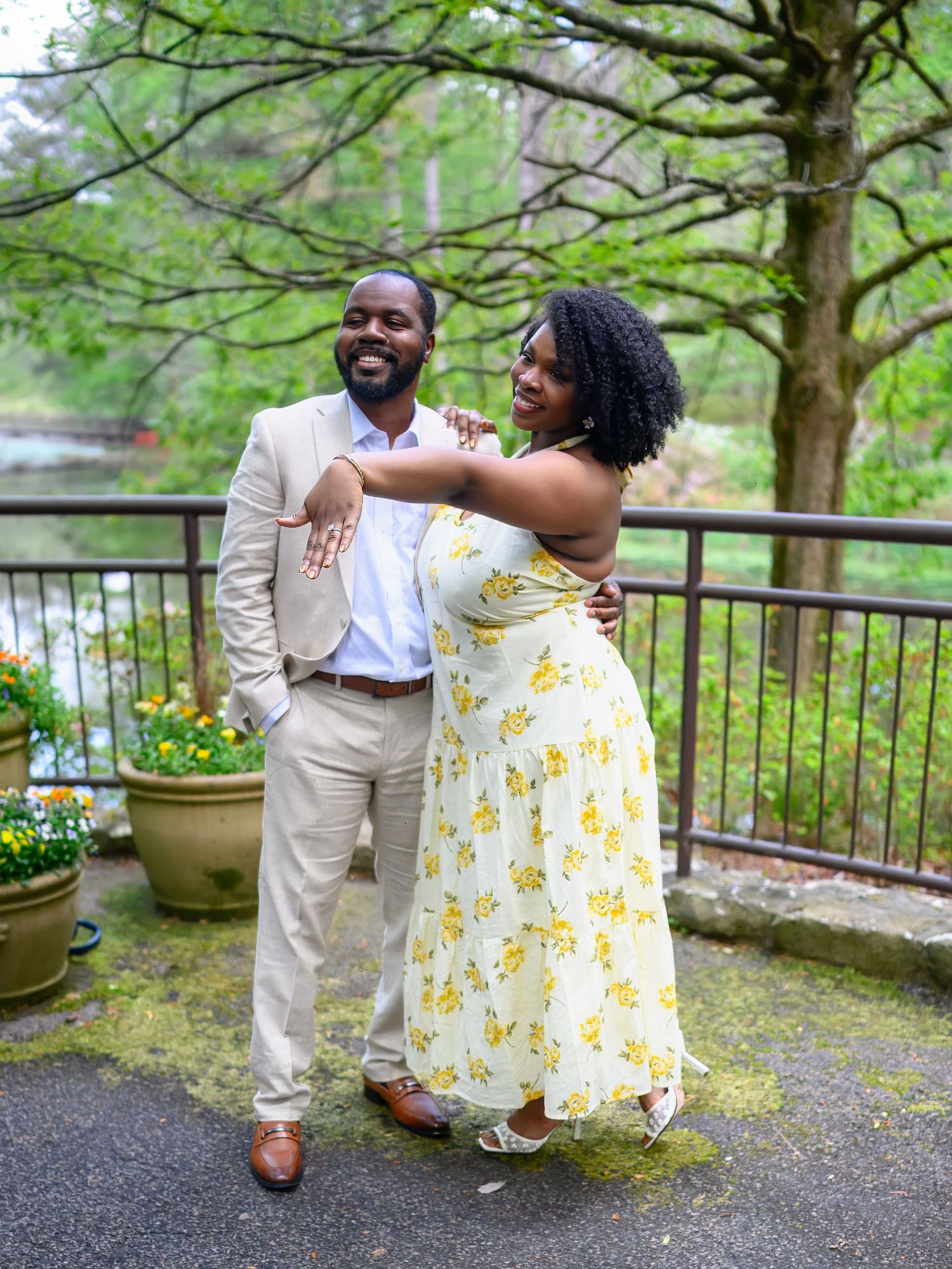 A smiling couple posing together outdoors in a garden, woman in yellow floral dress embracing man in tan suit.