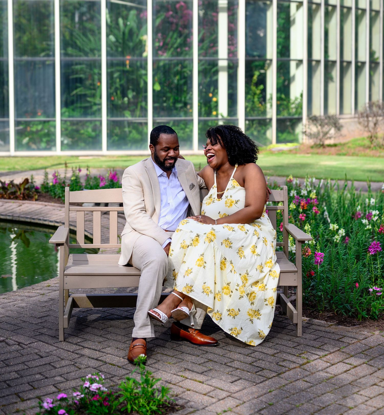Couple sitting together on a wooden bench laughing in a botanical garden with a greenhouse backdrop.