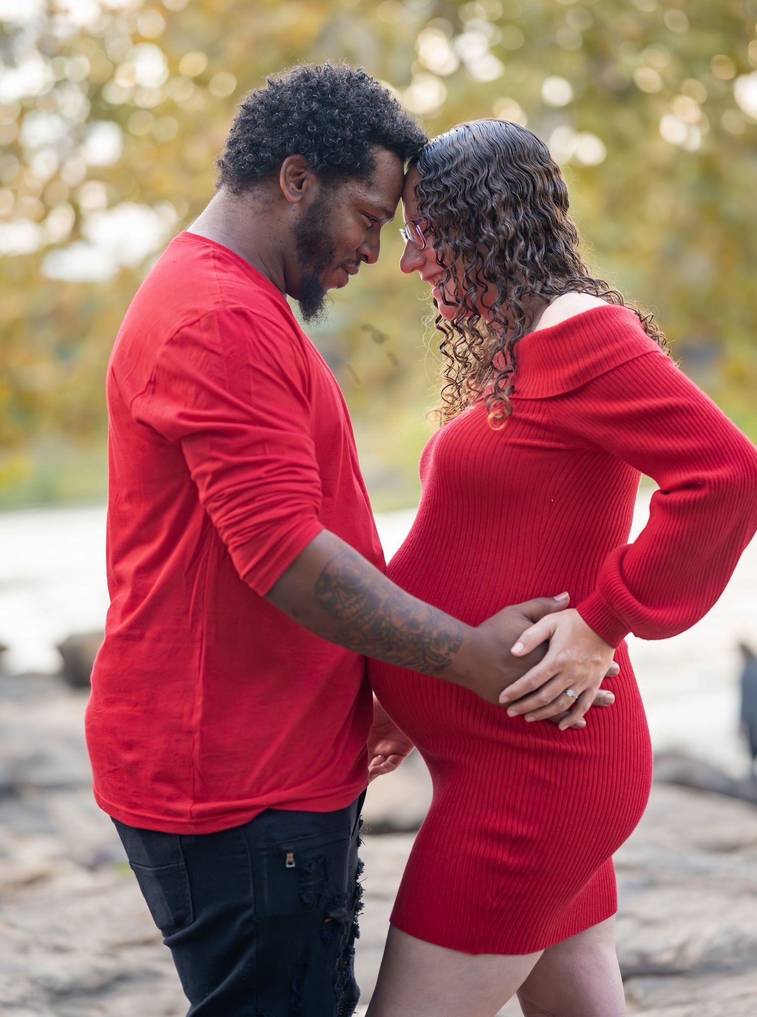 Candid maternity photos of family embracing on rocky riverbank in red outfits.