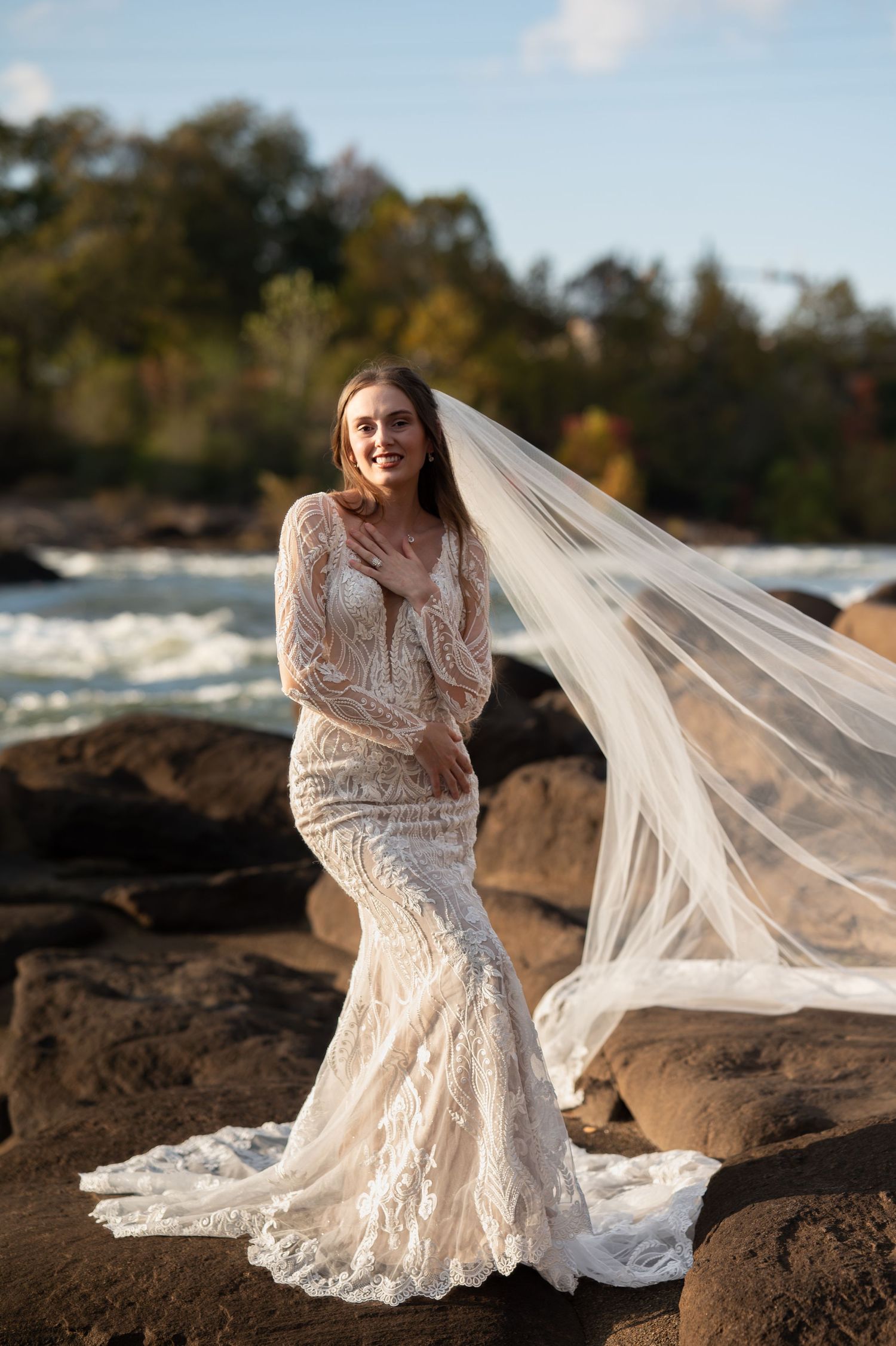 A bride's lace gown and veil catch the sunlight as she poses on rocky terrain near water.