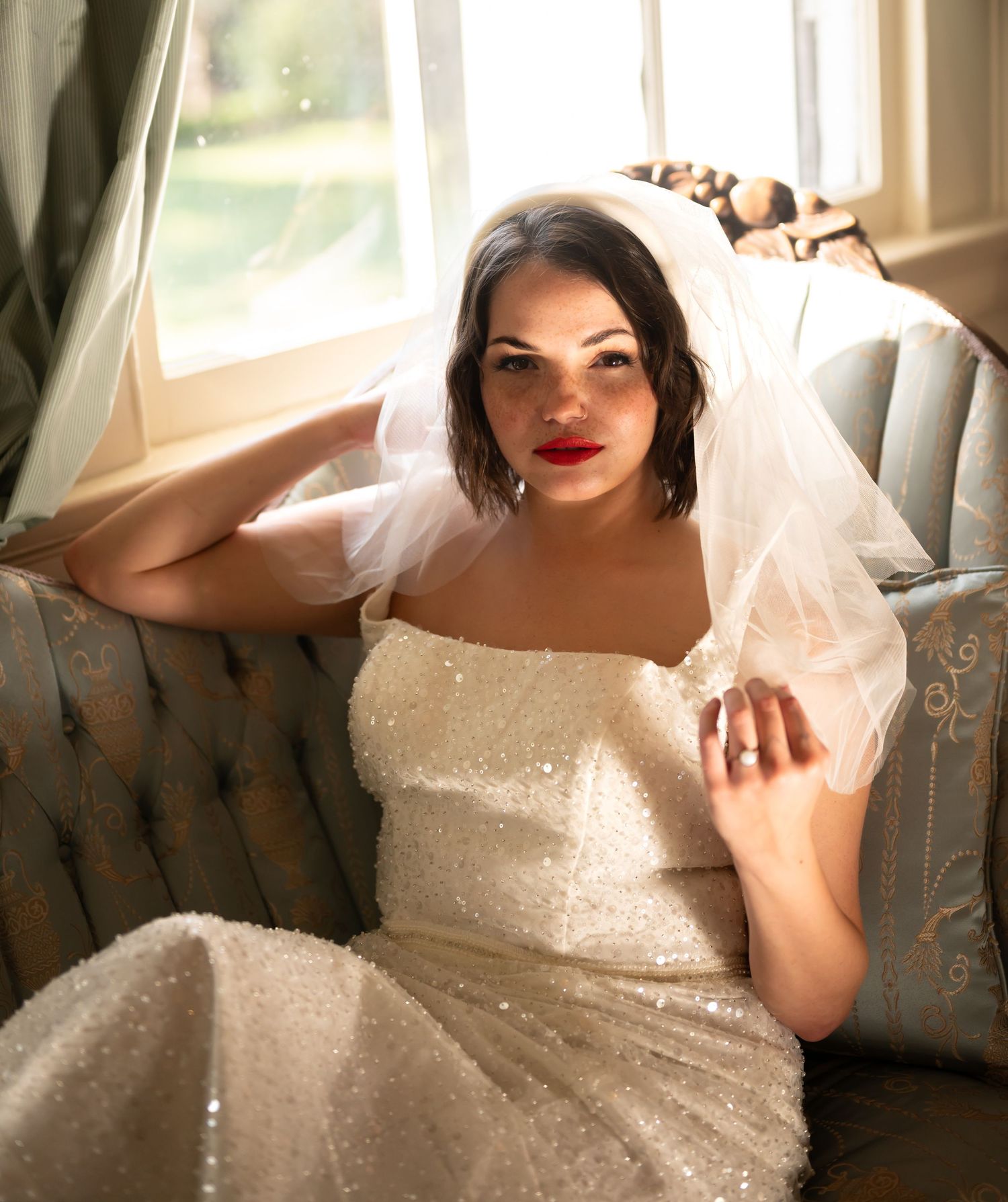 A person in a sparkly wedding dress with short sleeves sits elegantly on an upholstered chair near a window.