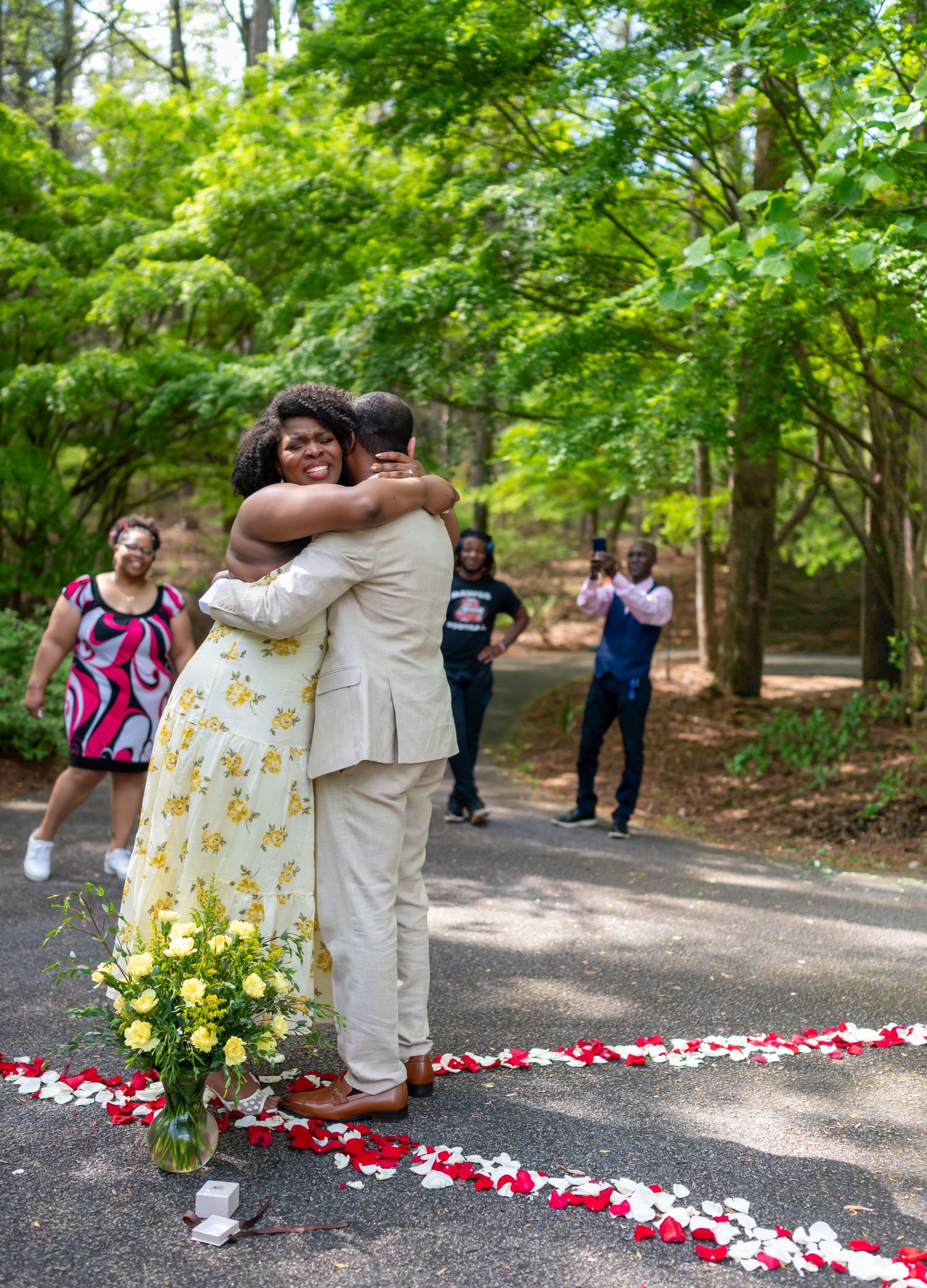 Couple embraces joyfully on a petal-lined path surrounded by lush green trees during an outdoor proposal.