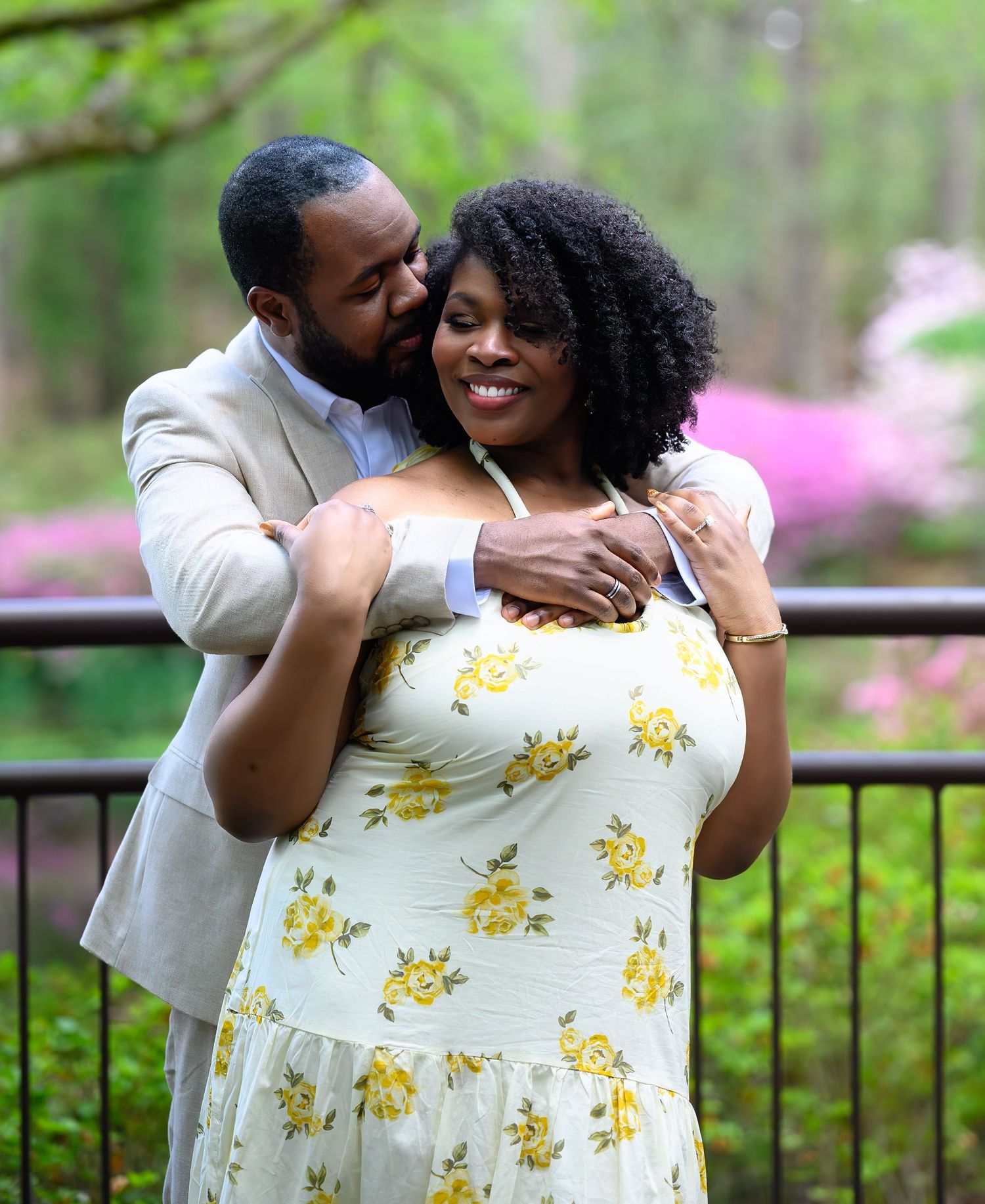 Man embracing woman from behind on a garden bridge surrounded by pink azaleas in bloom.