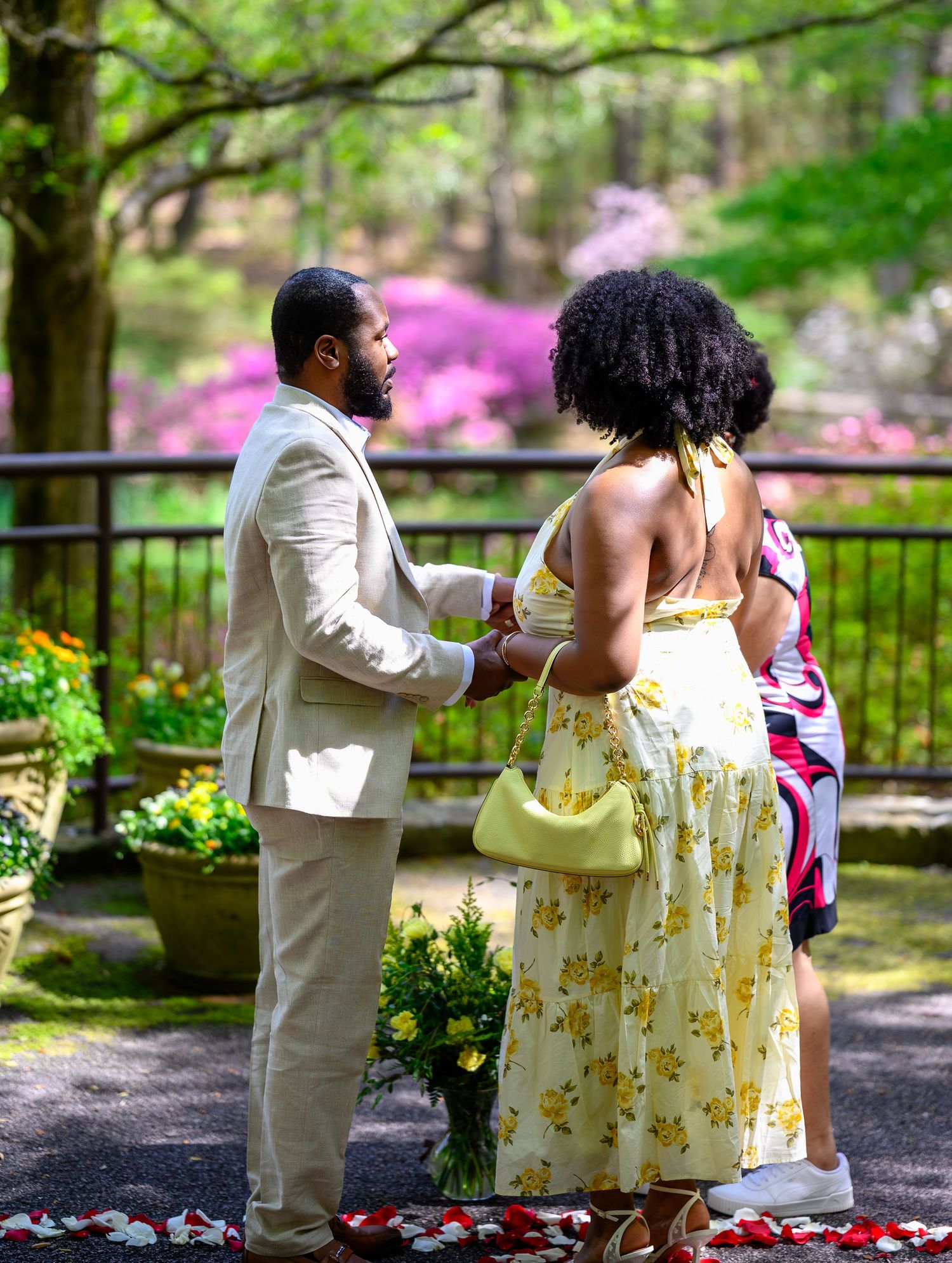 A man in a beige suit holds a woman's hands in a garden with colorful blooming flowers in the background.