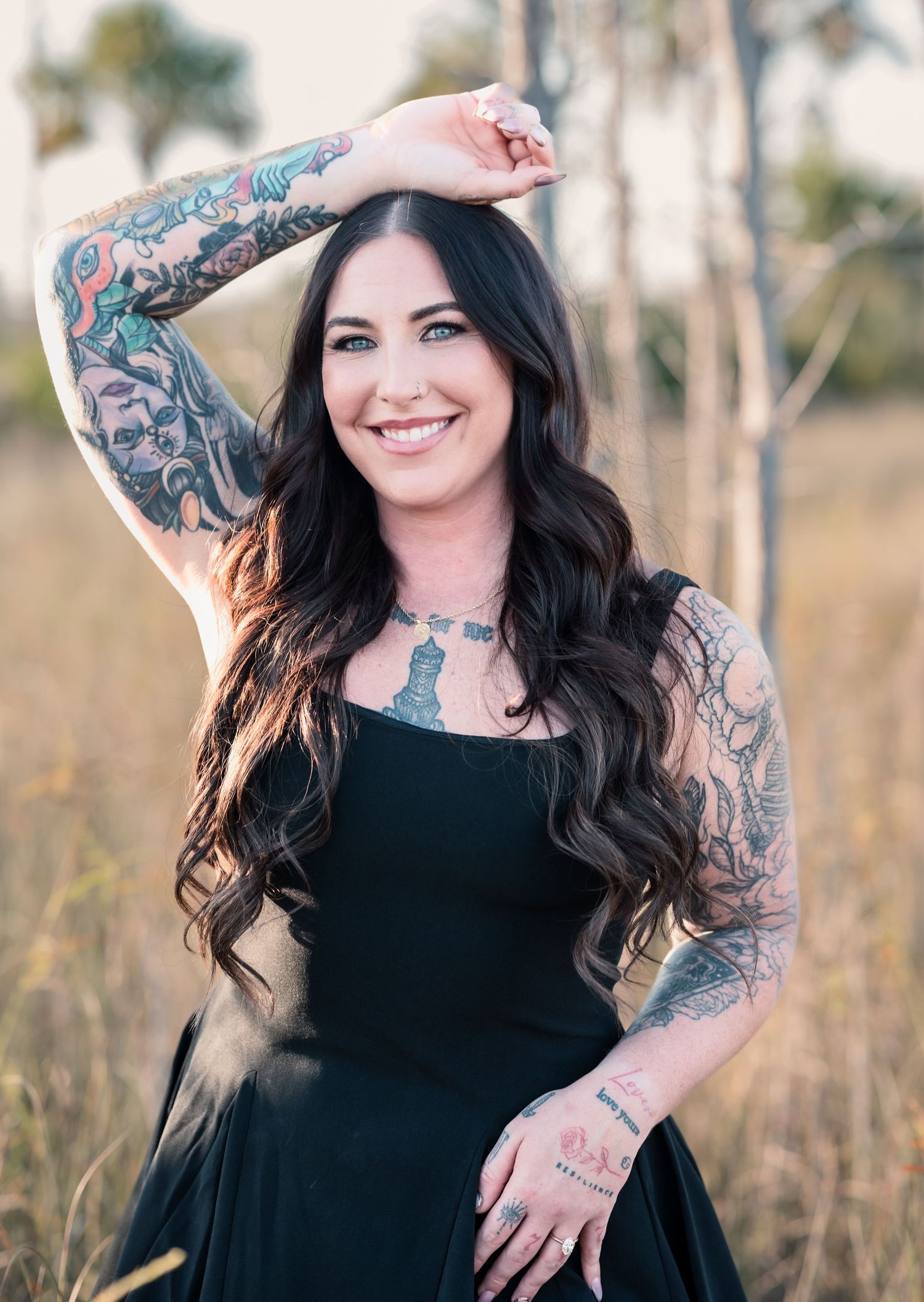 Woman with dark hair and tattoos poses confidently in black dress outdoors.