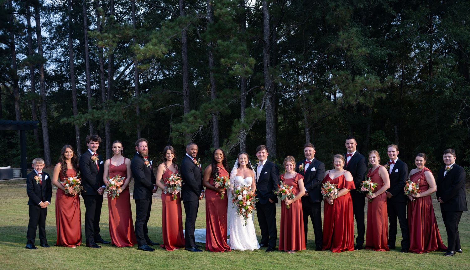 Wedding party in burgundy dresses and black suits poses outdoors among pine trees.