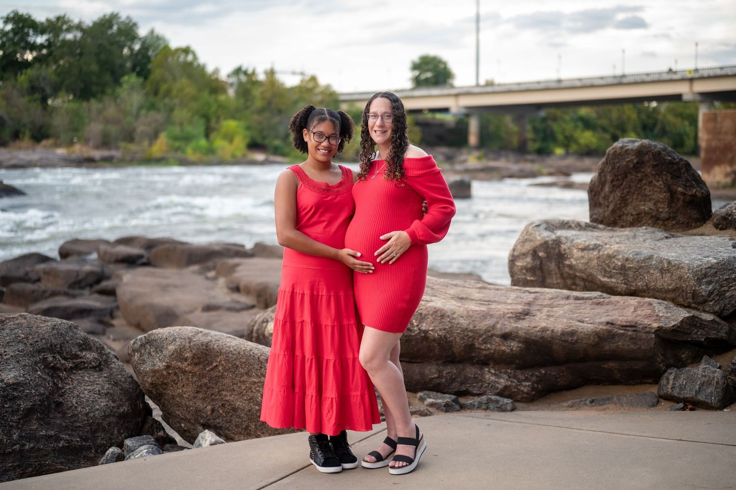 A couple in matching red dresses poses together on rocky coastal terrain during a sunset photoshoot.