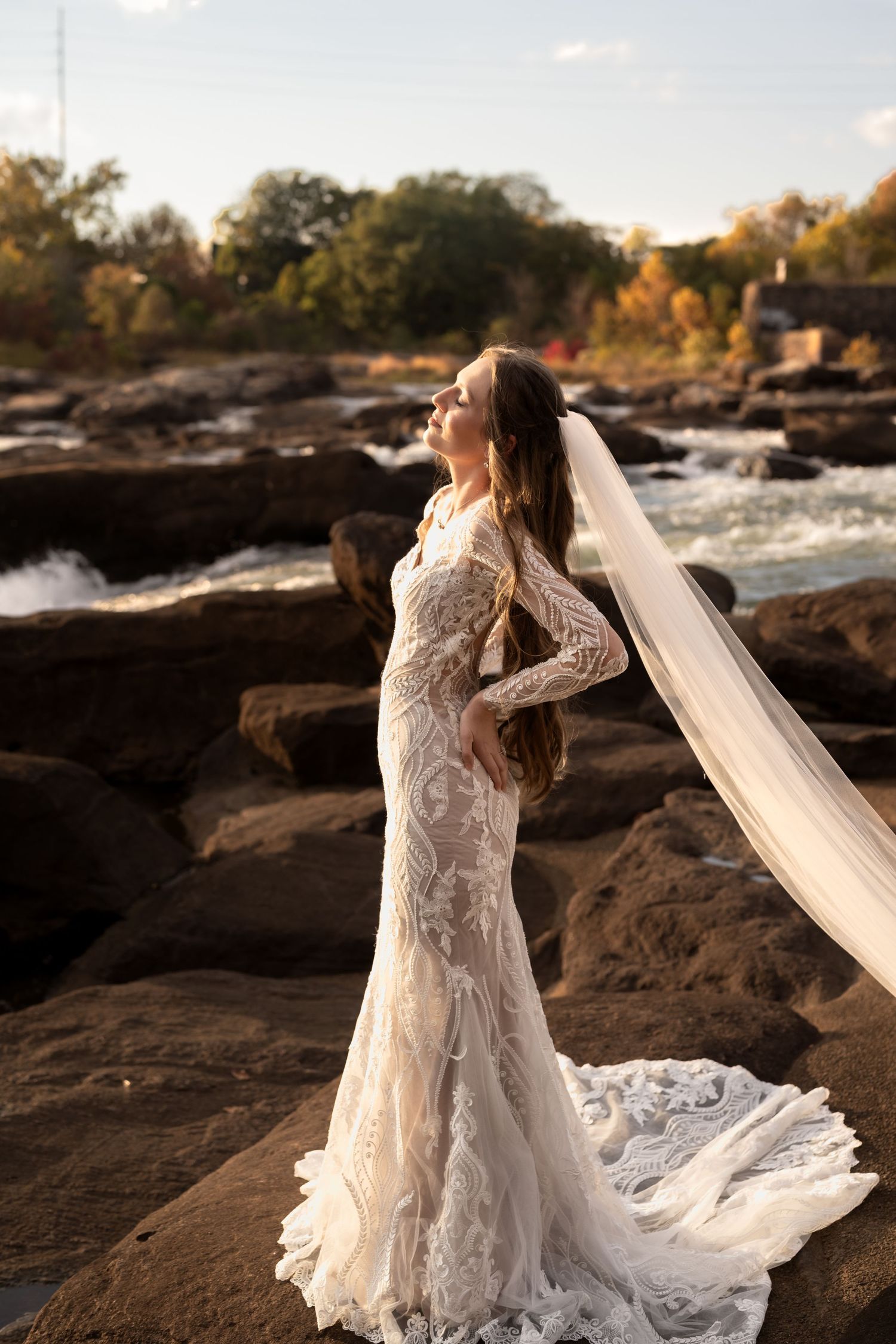 Dramatic bridal portraits on rocky riverbank with long-sleeved lace gown and cathedral veil.