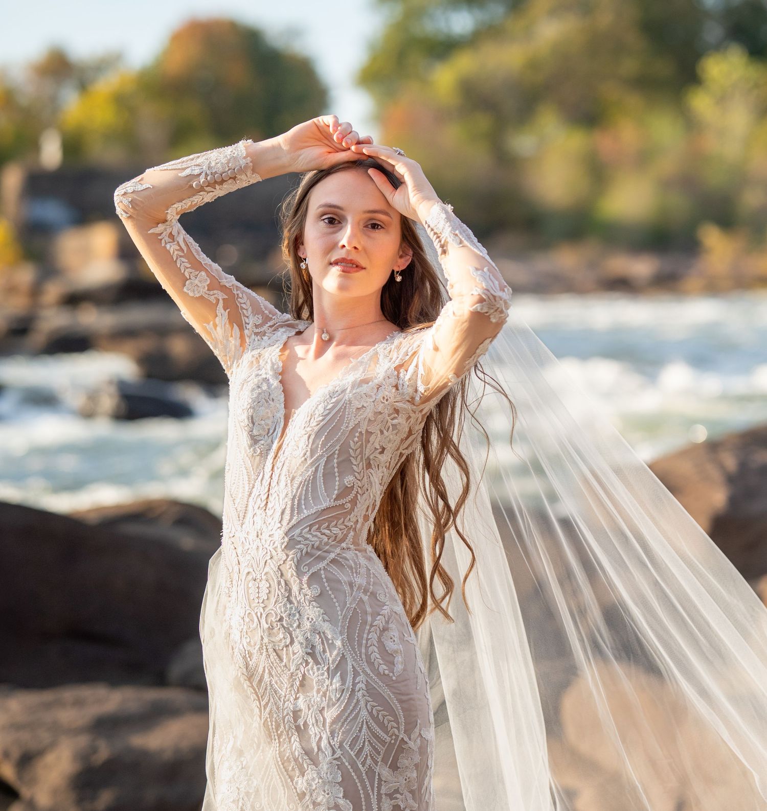 A bride in an ornate white gown stands on rocky terrain with flowing veil catching the golden sunlight.