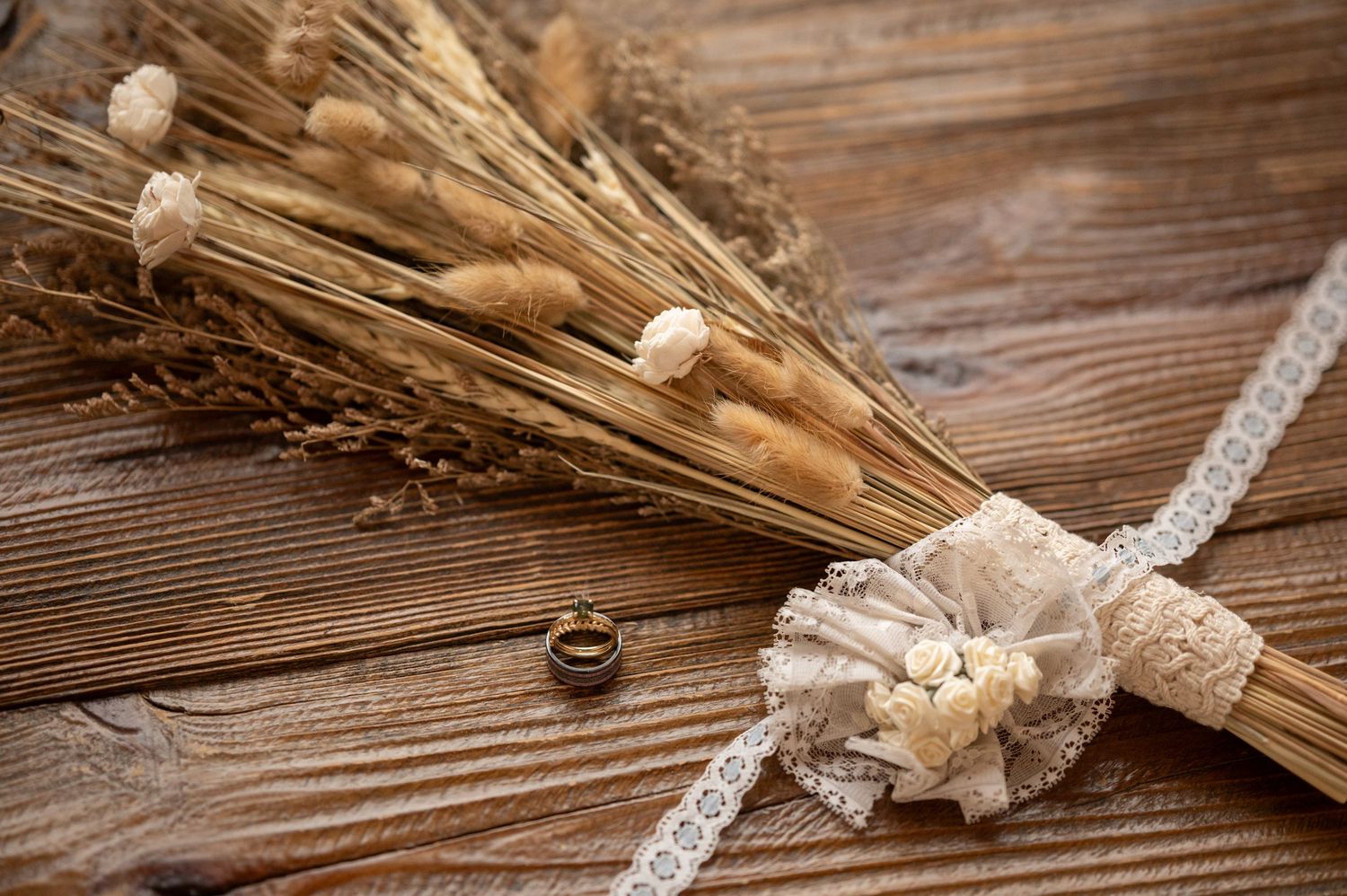 Wedding rings and lace details with dried wheat on wooden background.