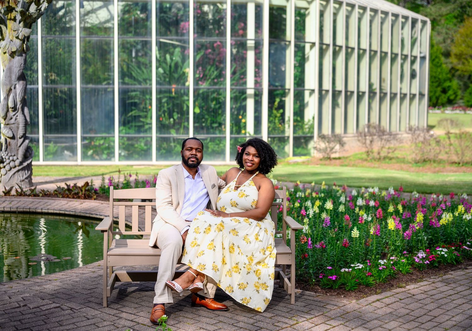 Engaged couple sitting on a garden bench near tulips and a greenhouse in coordinating spring outfits.