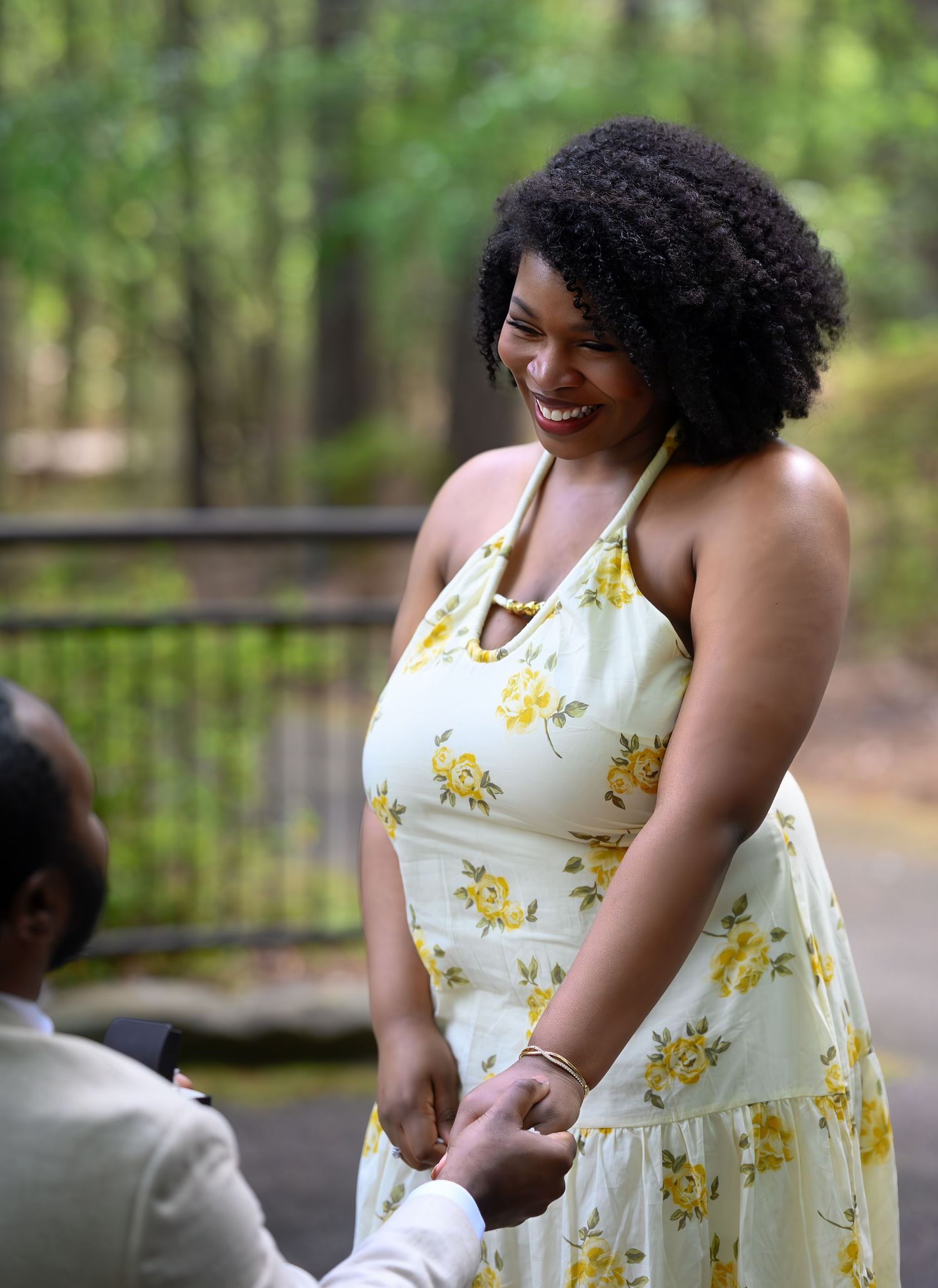 A joyful woman in a yellow floral dress smiles while holding hands during an outdoor marriage proposal in a wooded park.