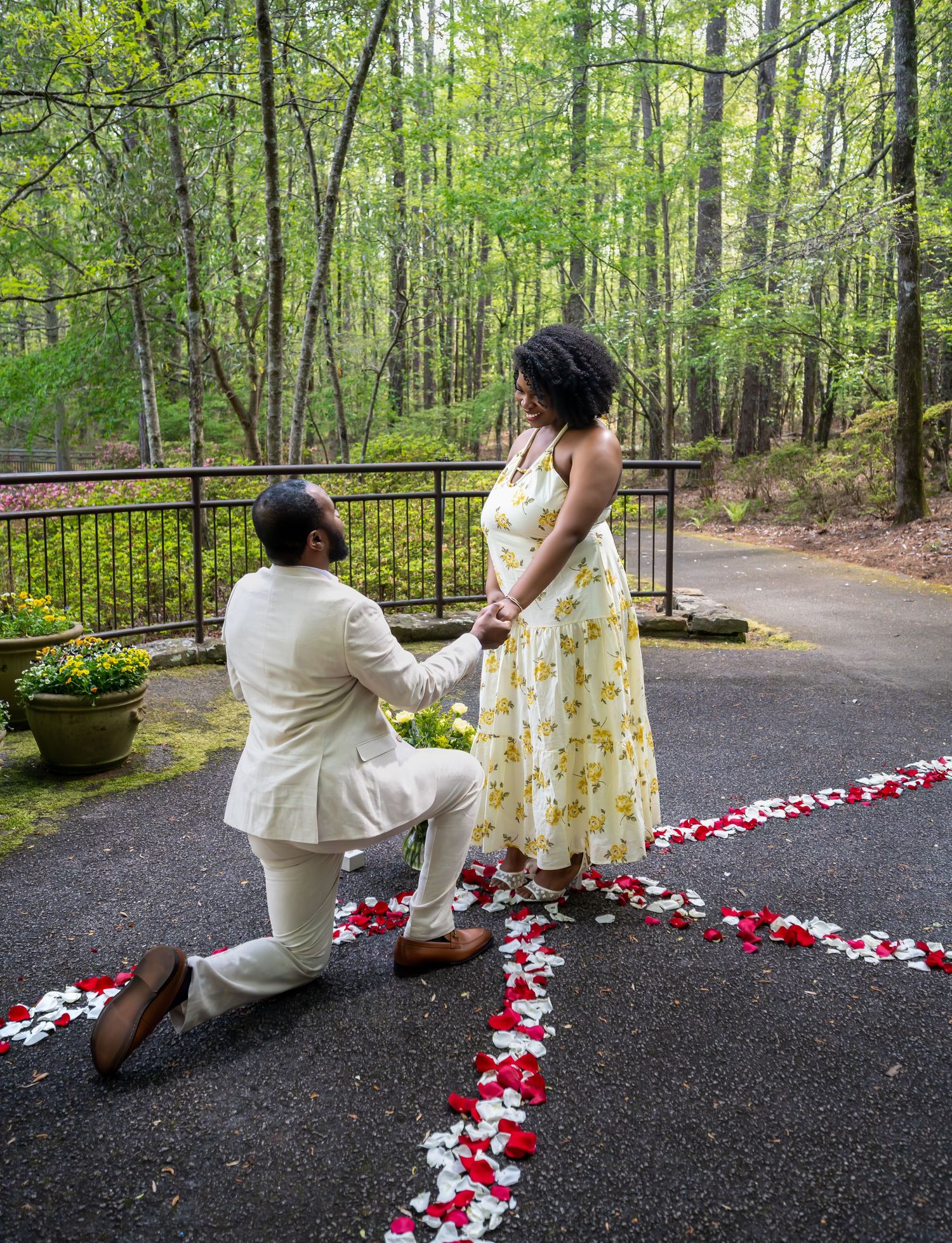 Man kneeling on one knee proposing to a woman in a yellow floral dress on a rose petal path.