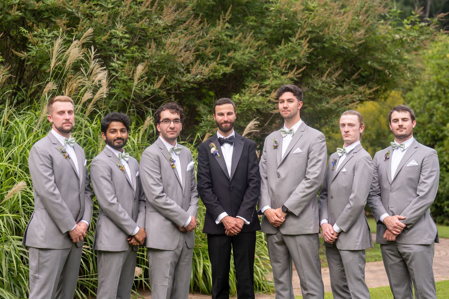 A wedding party of seven men in formal attire stands together outdoors with tall grass in the background.