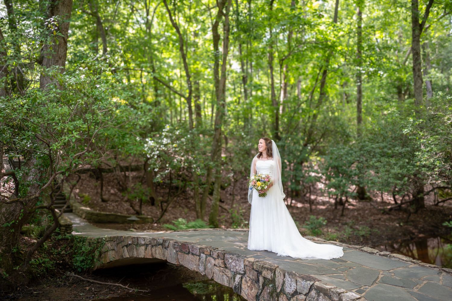 A bride in a white wedding dress poses on a rustic stone bridge surrounded by lush green forest scenery.