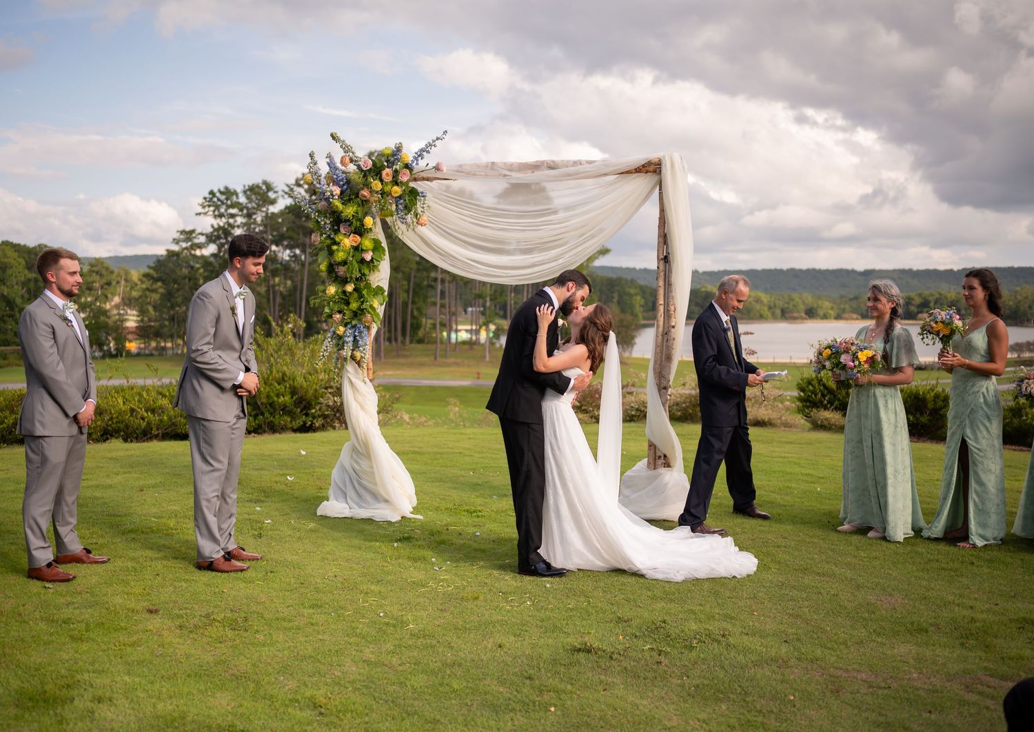 Wedding guests gather on a lush green lawn for an outdoor ceremony with white fabric draping and natural scenery.
