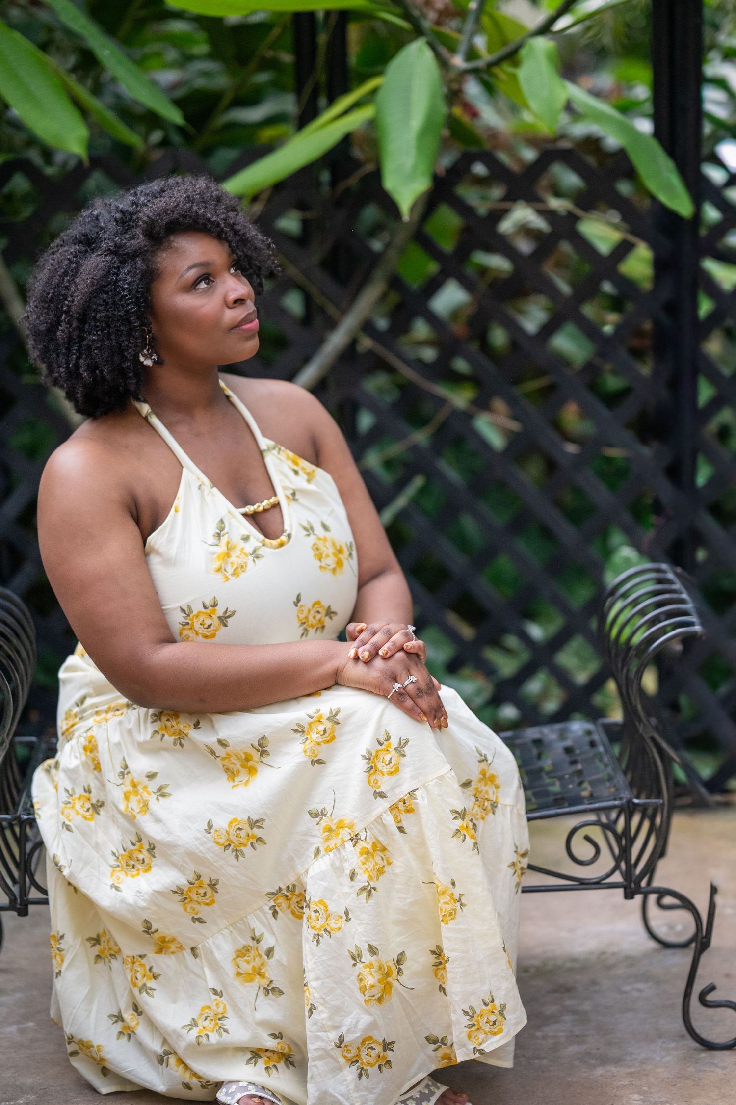 An elegant woman in a yellow floral halter dress sits gracefully on an ornate bench in a garden.