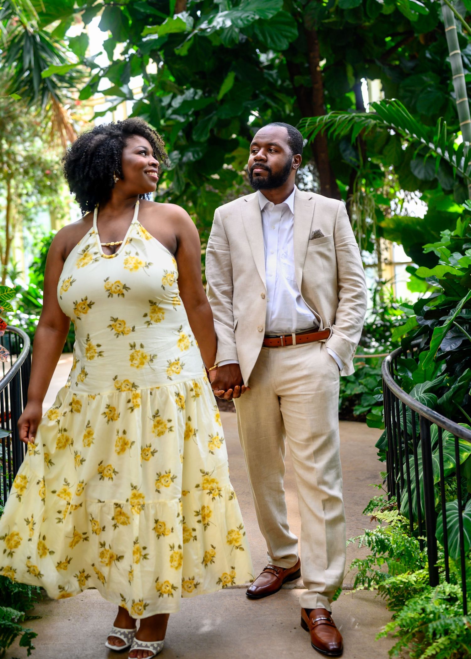 An elegant couple walks hand in hand through a tropical greenhouse, smiling at each other lovingly.