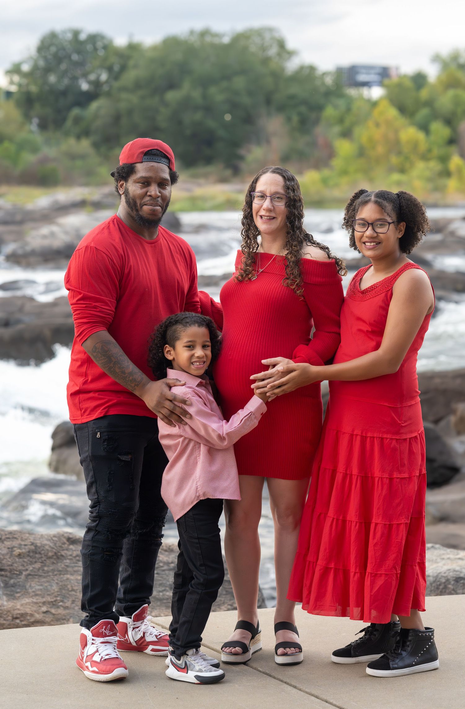 Family in coordinating red outfits poses together on rocky riverbank during maternity photoshoot.
